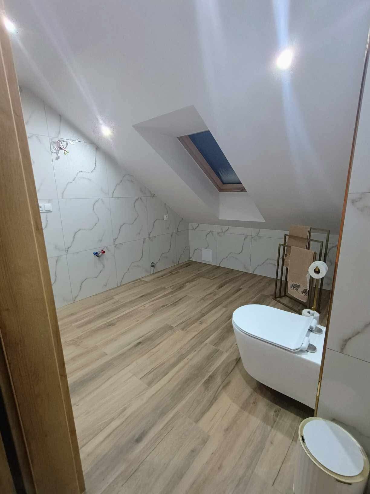 Nowoczesna łazienka na poddaszu z marmurowymi płytkami na ścianach, drewnopodobną podłogą, toaletą wiszącą i oknem dachowym