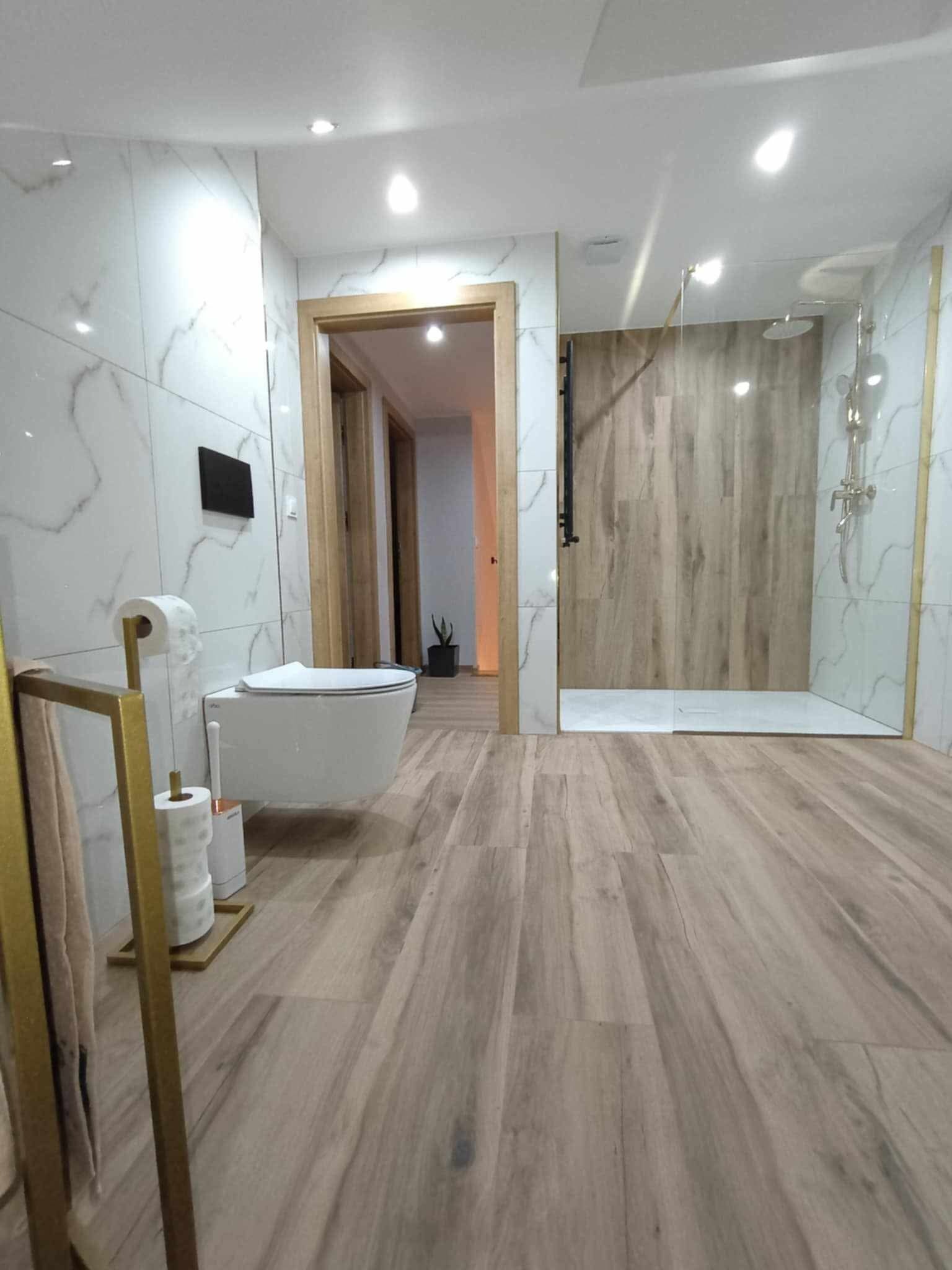 Nowoczesna łazienka z kabiną prysznicową typu walk-in, podwieszaną toaletą i drewnopodobną podłogą, wykończona białymi płytkami imitującymi marmur.