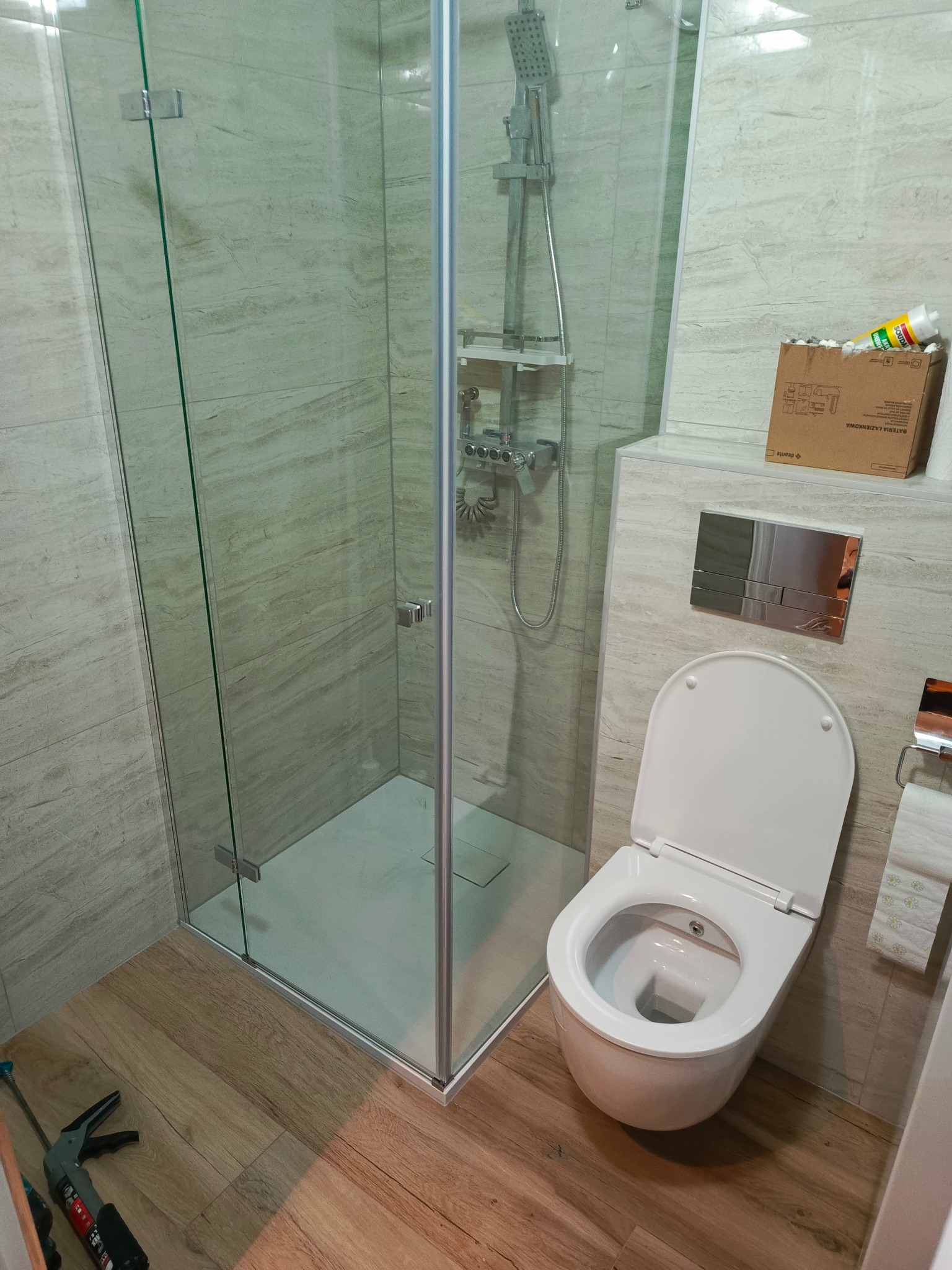 Nowoczesna łazienka z kabiną prysznicową typu walk-in, wiszącą toaletą i drewnopodobną podłogą, widoczny karton po baterii łazienkowej i narzędzie do silikonu.