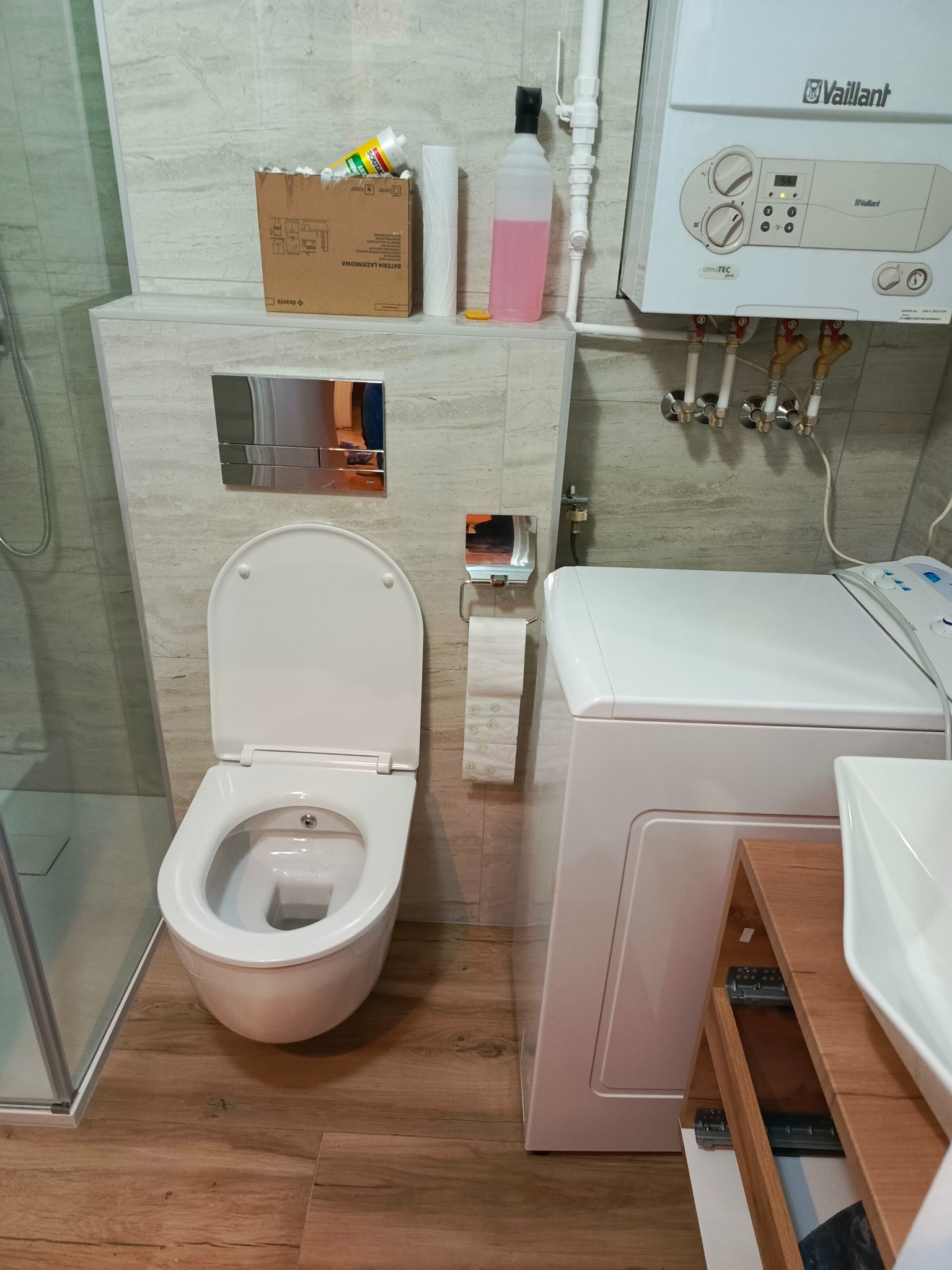 Nowoczesna łazienka z podwieszaną toaletą, pralką, umywalką na drewnianym stelażu oraz bojlerem Vaillant na ścianie, widoczne akcesoria łazienkowe i karton po materiałach budowlanych.