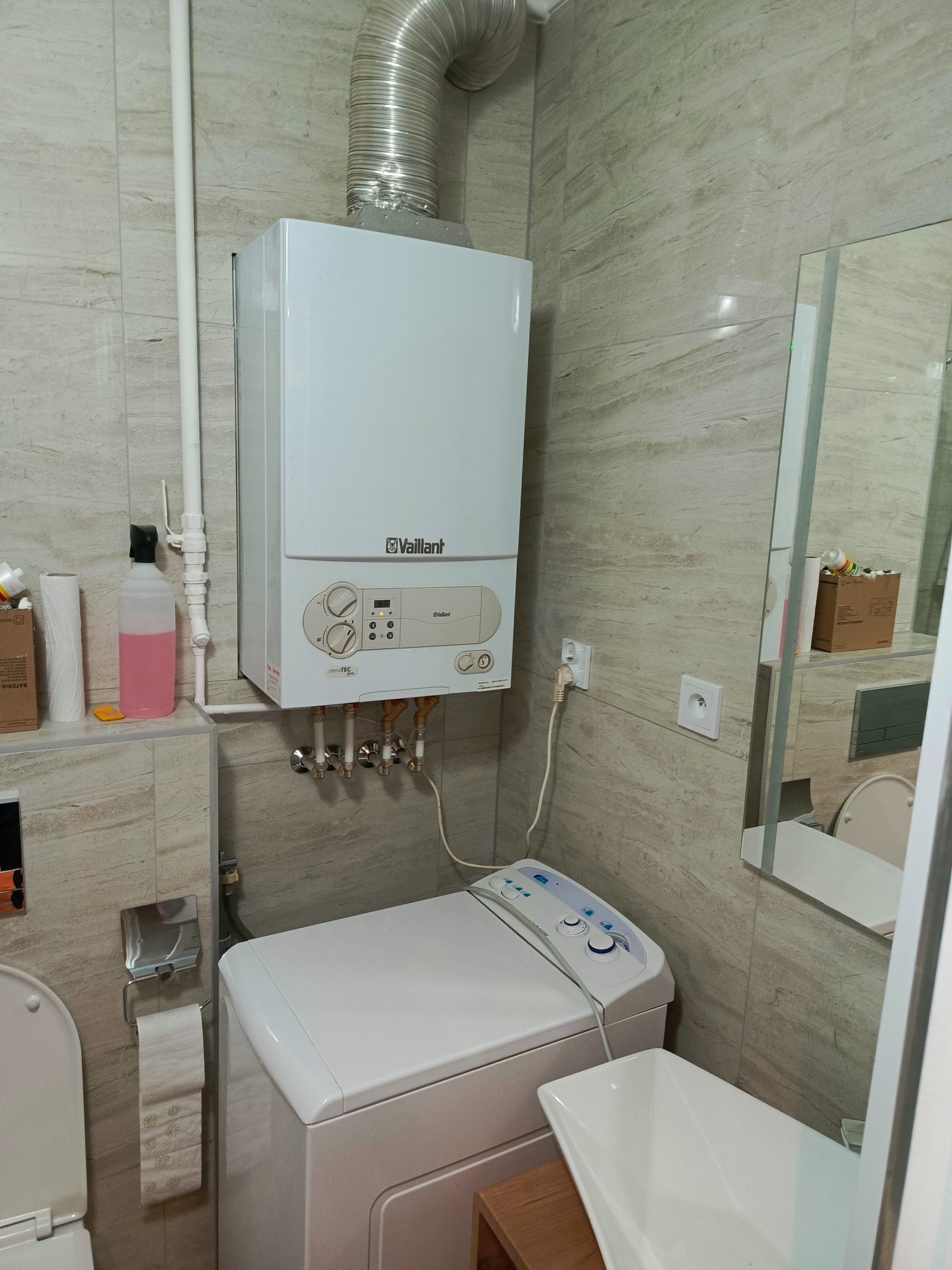 Kompaktowa łazienka w Siedliskach z piecem gazowym Vaillant nad pralką, widoczne beżowe płytki imitujące kamień i fragment umywalki.