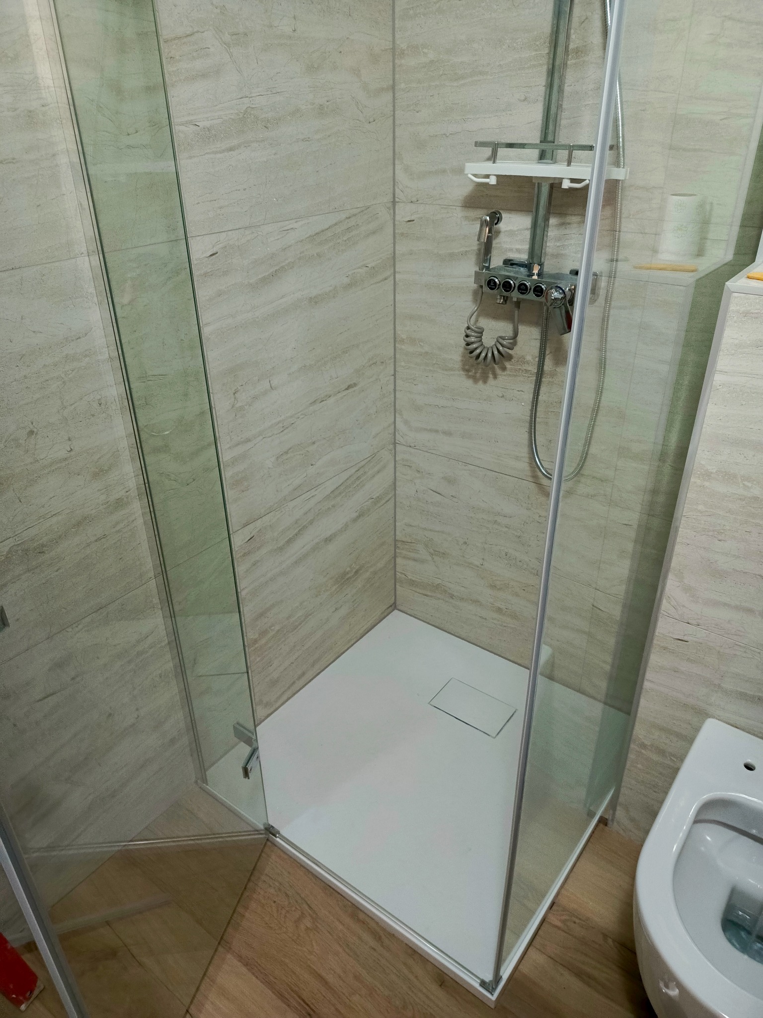 Kabina prysznicowa z transparentnego szkła, biały brodzik z odpływem liniowym, bateria natryskowa z półką i słuchawką, płytki ścienne imitujące trawertyn, podłoga z paneli drewnopodobnych, fragment...