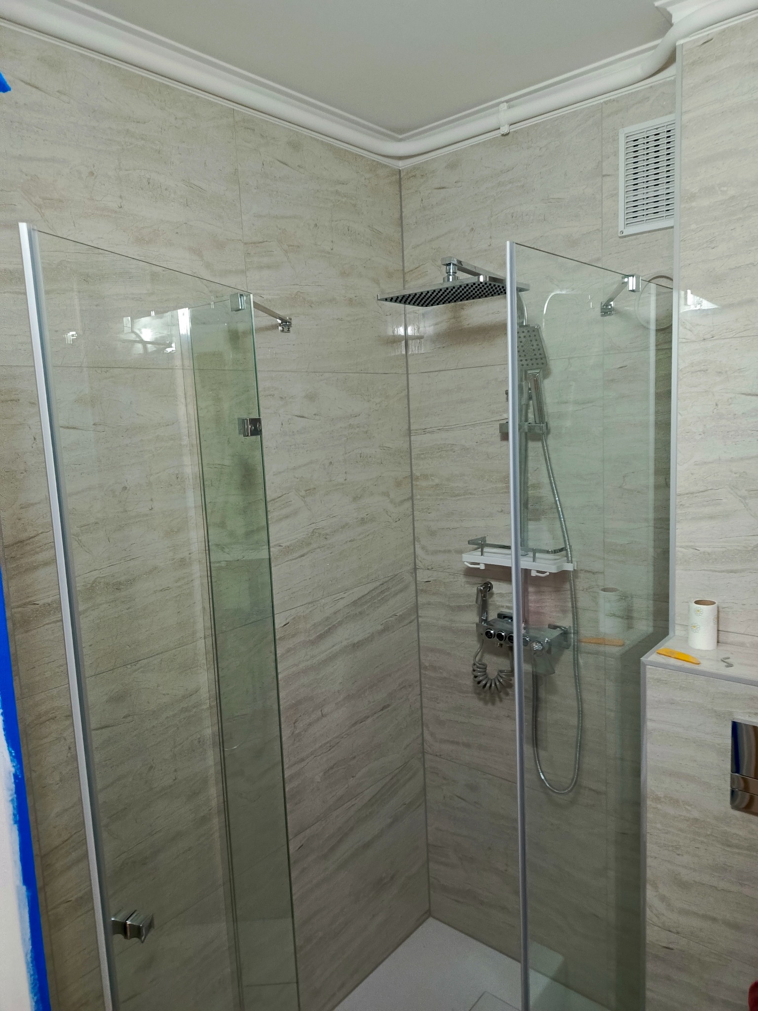 Wykończona kabina prysznicowa typu walk-in z deszczownicą i panelem prysznicowym, wyłożona płytkami imitującymi kamień w odcieniach beżu i szarości, z widoczną kratką wentylacyjną i akcesoriami...