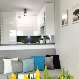 Dorota Suchomska Nieruchomości w Dobrym Stylu - Jasne wnętrze mieszkania z aneksem kuchennym, sofa z poduszkami w odcieniach szarości i błękitu, na pierwszym planie bukiet żółtych tulipanów.