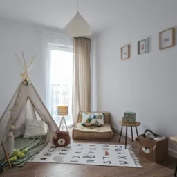 Jasny pokój dziecięcy z wigwamem ozdobionym kulkami, materacem pod oknem, dywanem z literami alfabetu i zwierzętami, abakusem oraz dekoracjami w kształcie misia.