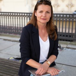 Dorota Suchomska Nieruchomości w Dobrym Stylu - Kobieta siedzi na ławce z laptopem, ubrana w marynarkę i dżinsy, w tle ozdobna balustrada i budynek.