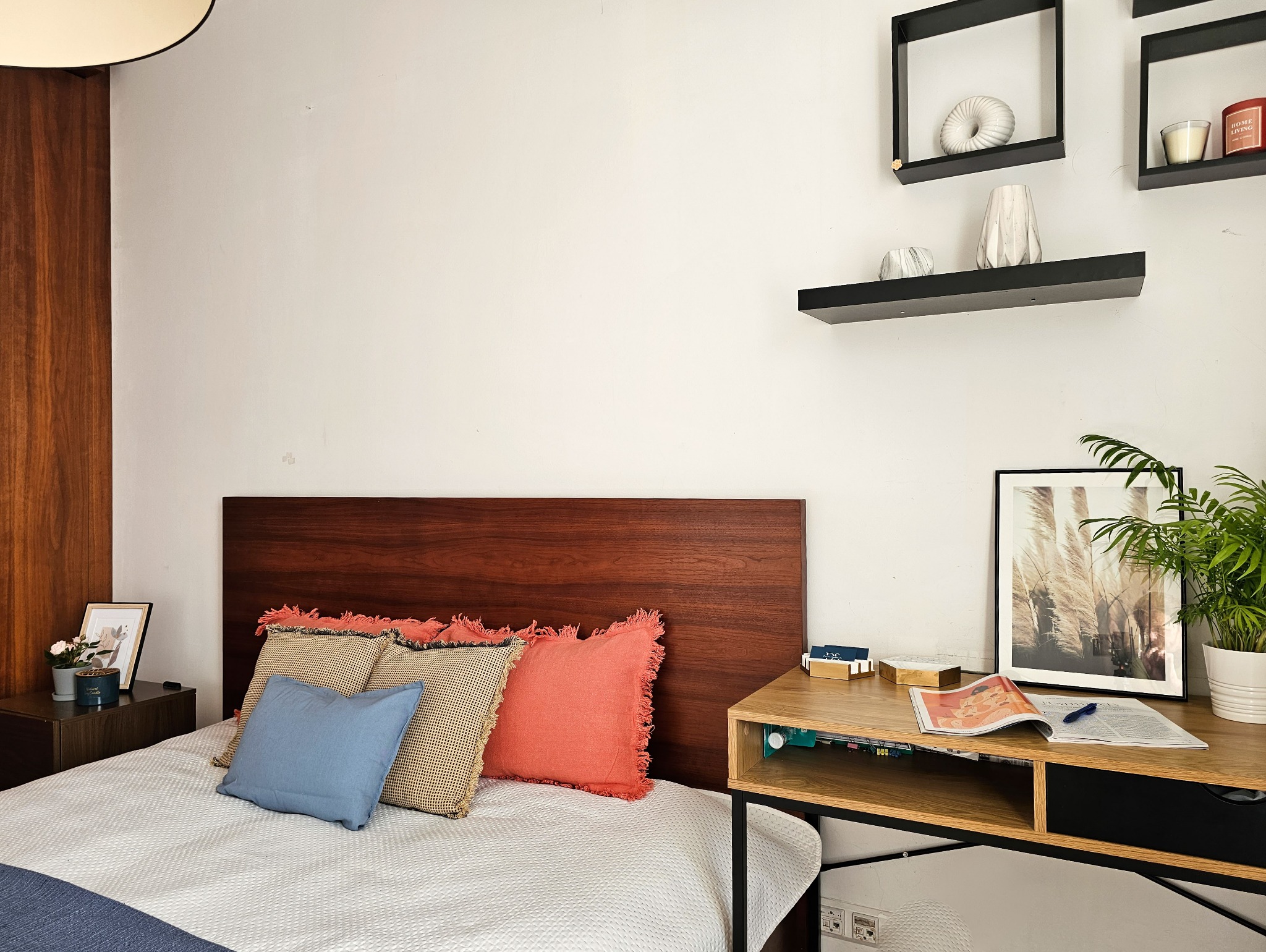 Nowoczesna sypialnia z drewnianym wezgłowiem, kolorowymi poduszkami, biurkiem z rośliną doniczkową i dekoracyjnymi półkami na ścianie.