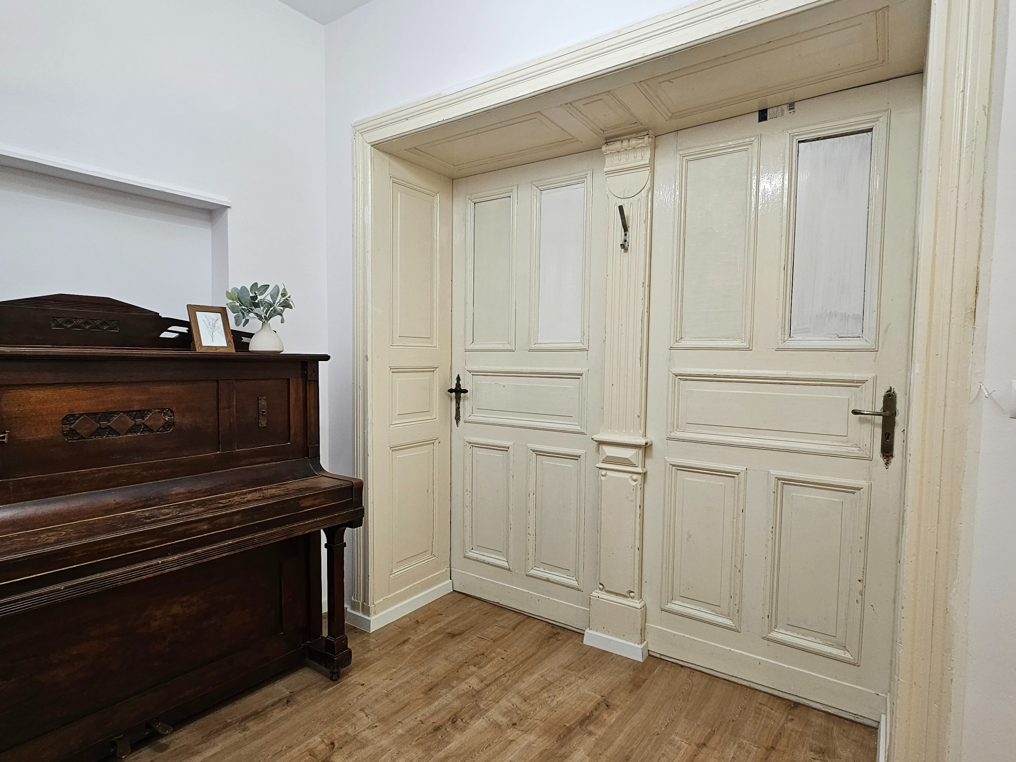 Wnętrze pokoju z odnowionymi, dwuskrzydłowymi drzwiami w stylu vintage, pianinem z ciemnego drewna i dekoracjami na nim.