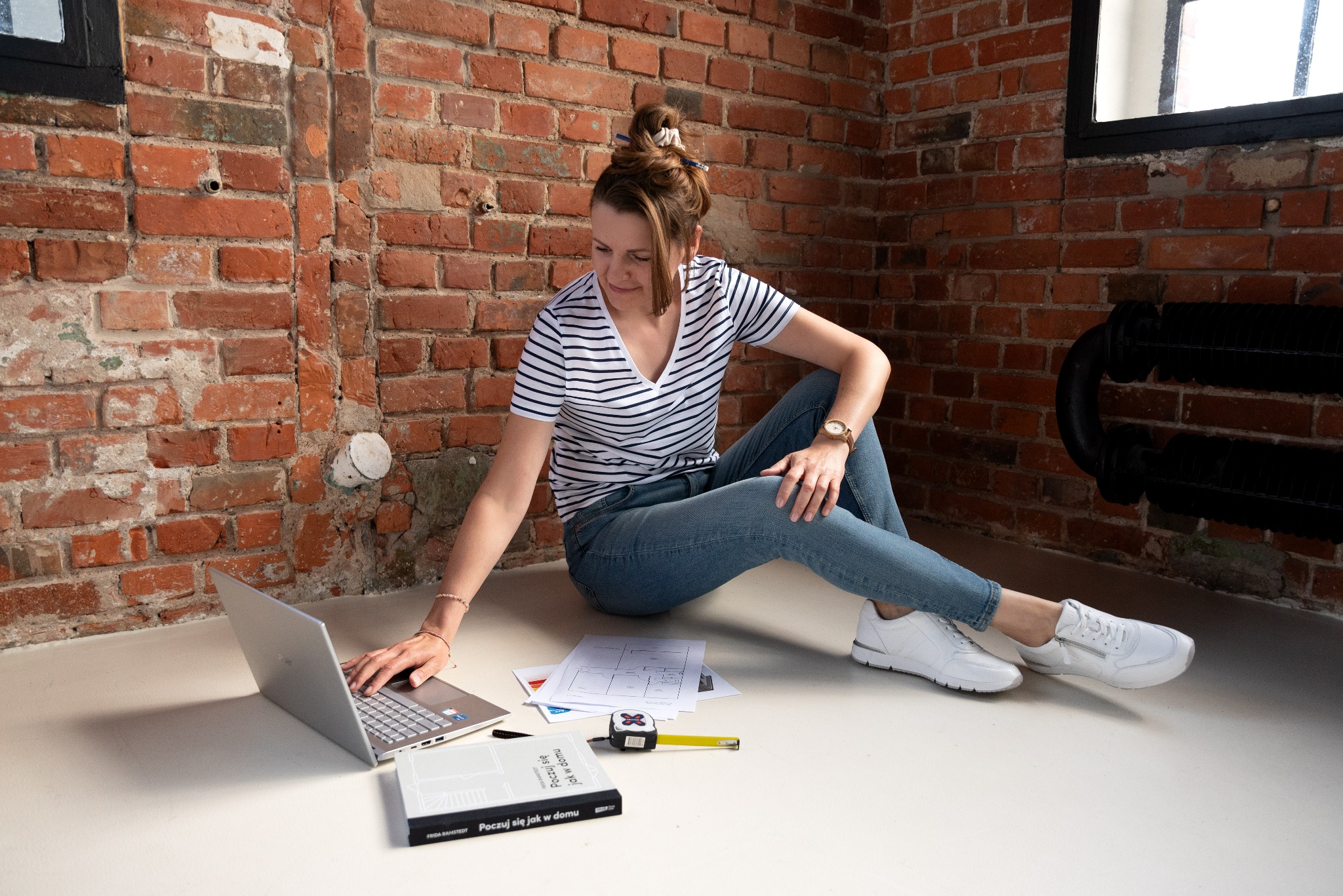 Kobieta siedzi na podłodze z laptopem, planami mieszkania i miarką, na tle ceglanej ściany z widocznymi rurami grzewczymi.