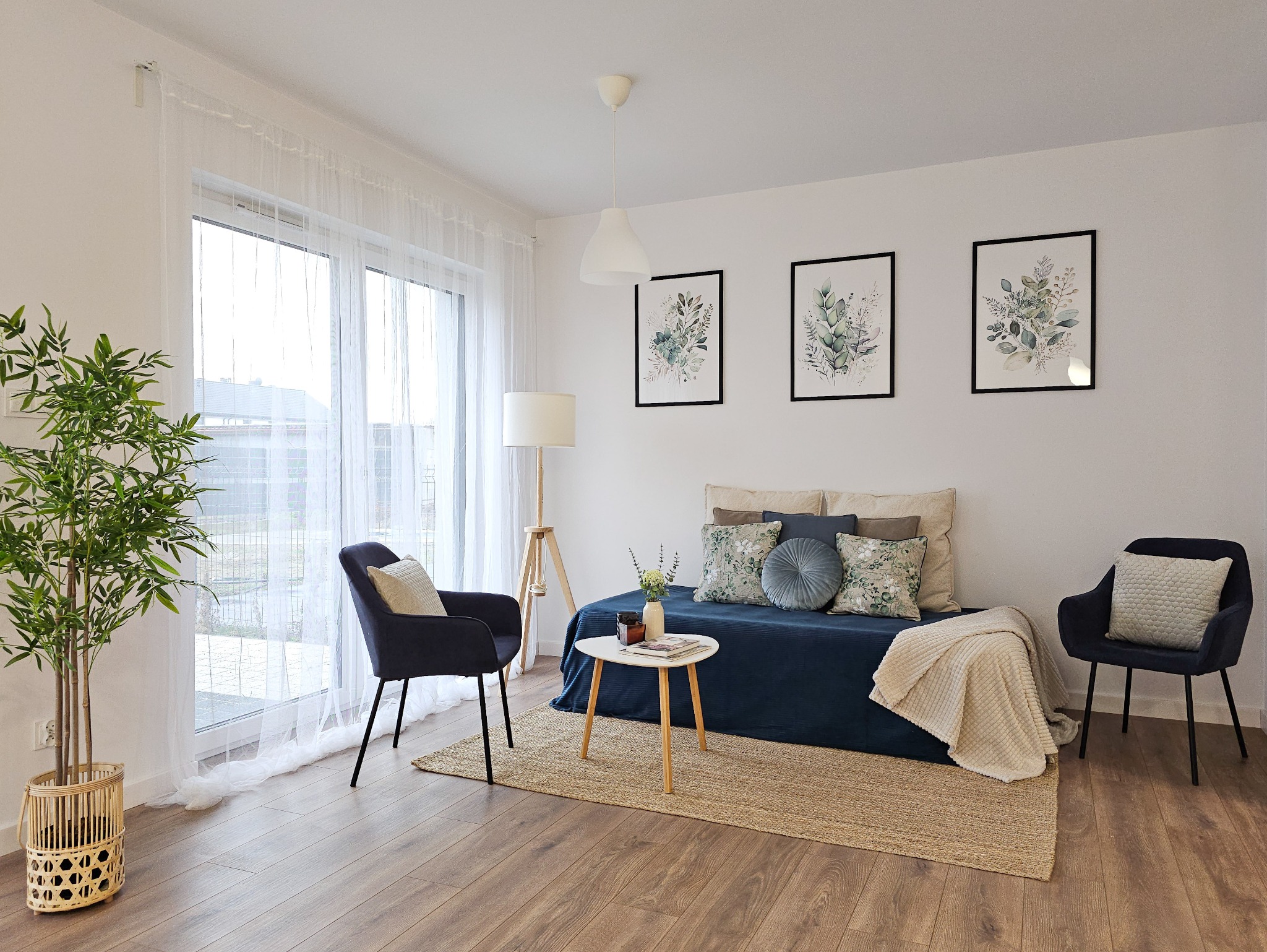 Jasny salon z niebieską kanapą, dwoma fotelami, stolikiem kawowym, rośliną w plecionym koszu i trzema obrazkami z motywami roślinnymi na ścianie.