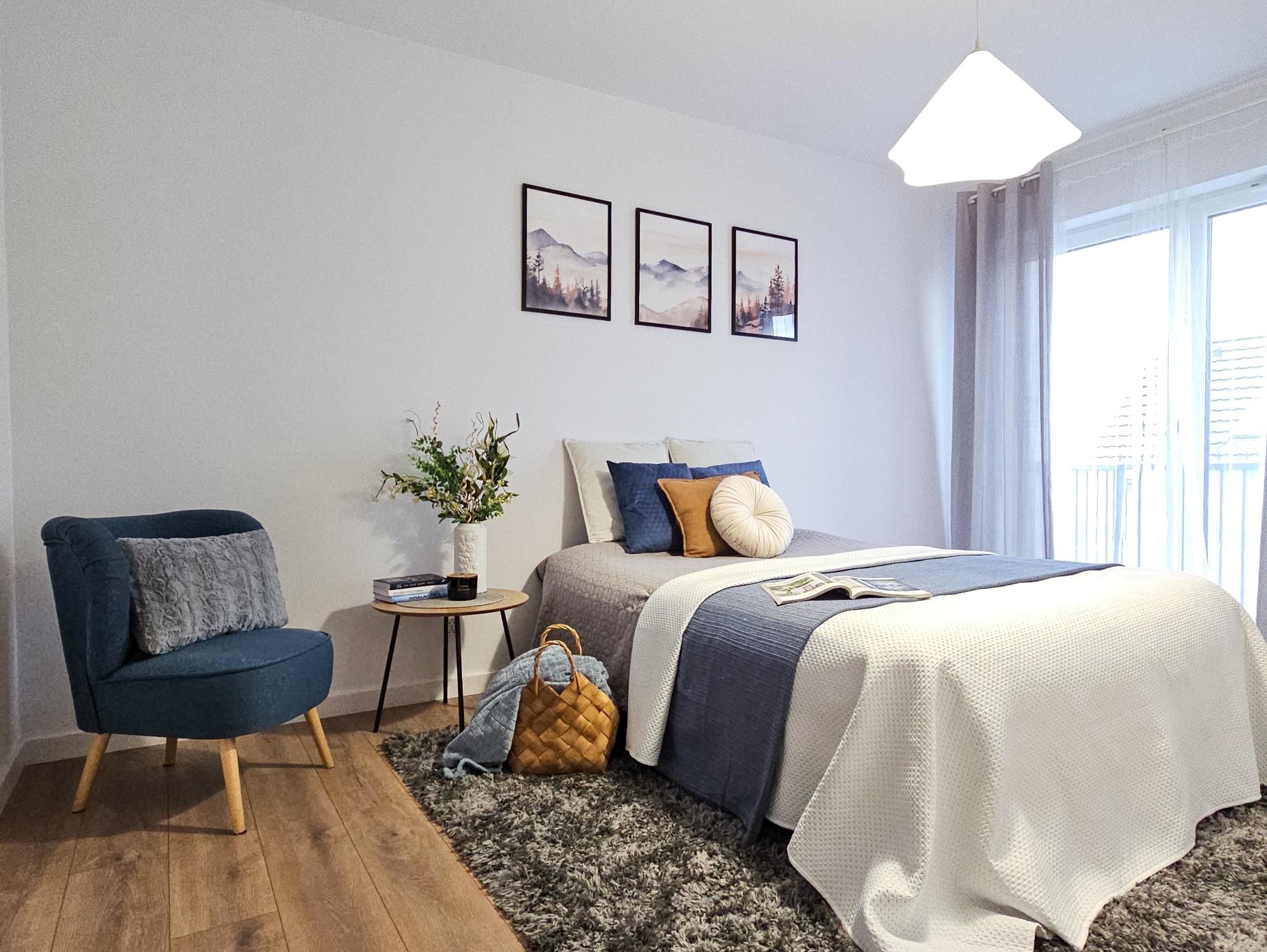 Aranżacja sypialni we Wrocławiu: Pokój z łóżkiem, krzesłem, stolikiem i trzema obrazami górskimi na ścianie. Na łóżku leżą poduszki i książka, a obok stoi wazon z kwiatami.