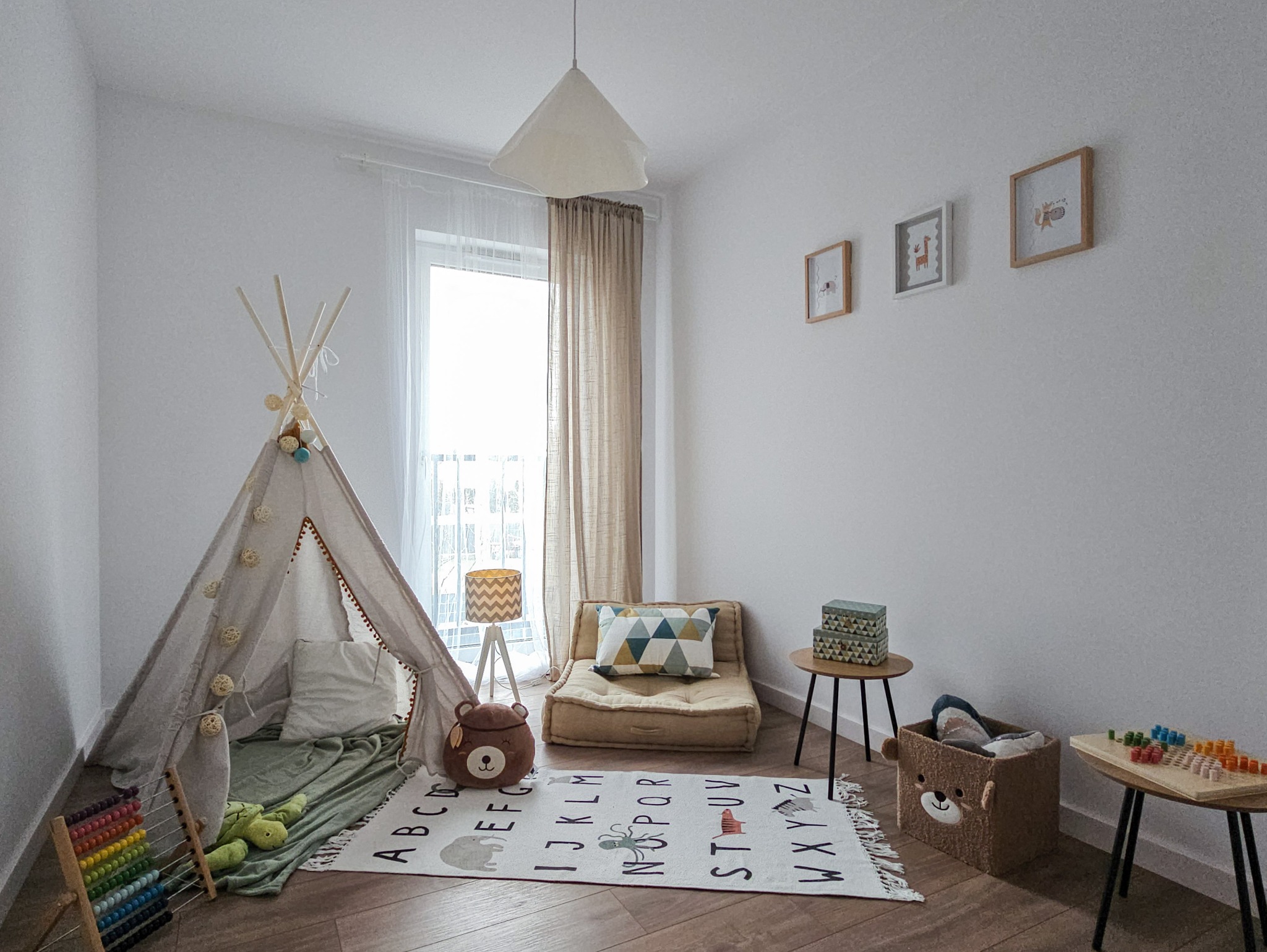 Jasny pokój dziecięcy z wigwamem ozdobionym kulkami, materacem pod oknem, dywanem z literami alfabetu i zwierzętami, abakusem oraz dekoracjami w kształcie misia.