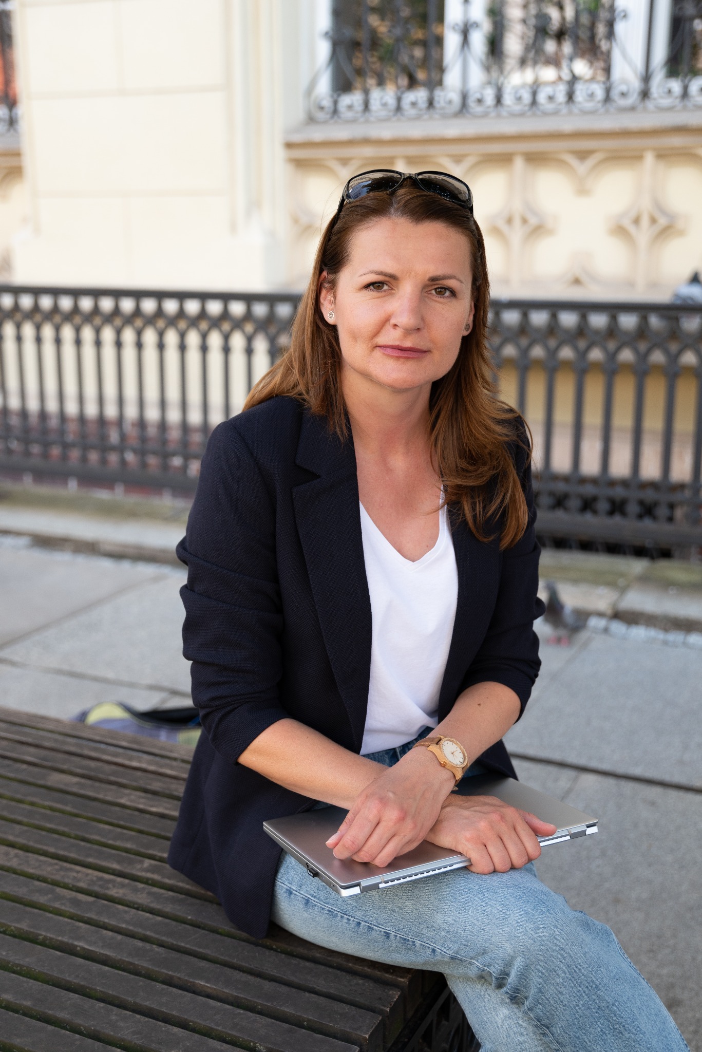 Kobieta siedzi na ławce z laptopem, ubrana w marynarkę i dżinsy, w tle ozdobna balustrada i budynek.