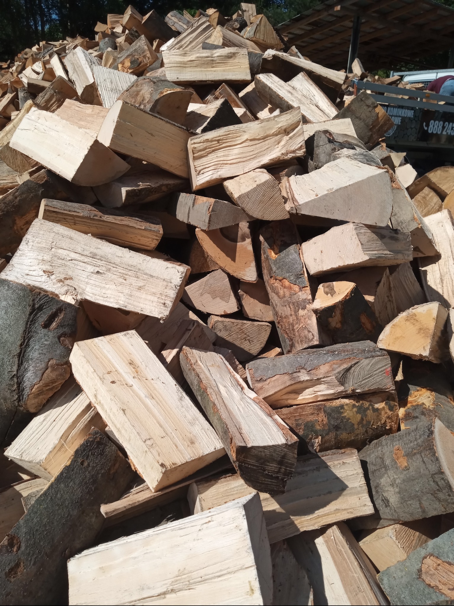 Stos pociętego drewna opałowego z widoczną przyczepką i numerem telefonu w tle.