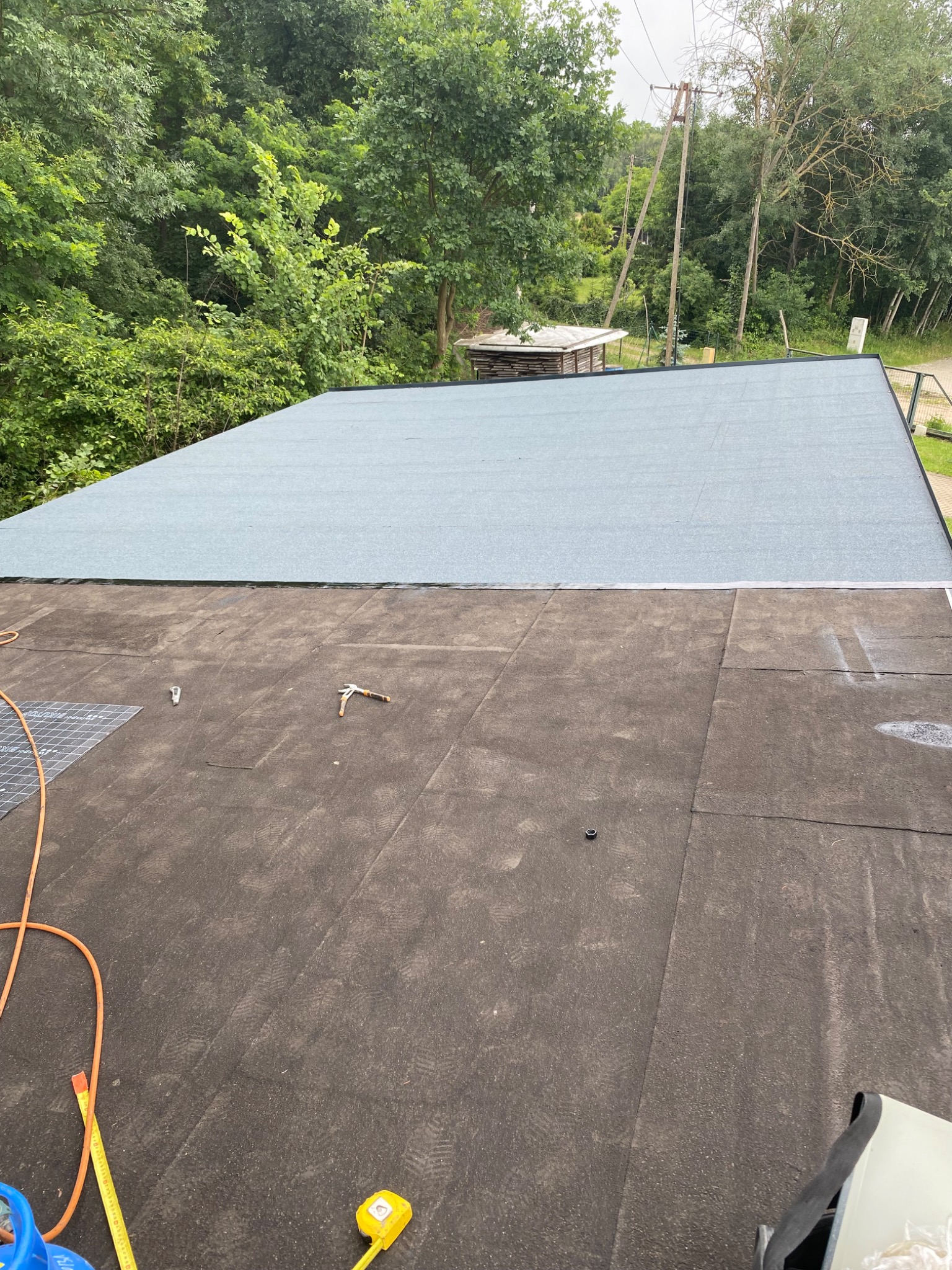 W trakcie prac dekarskich: częściowo pokryty papą dach płaski z widocznym narzędziem dekarskim, zwiniętą miarką i butlą gazową w rogu zdjęcia.