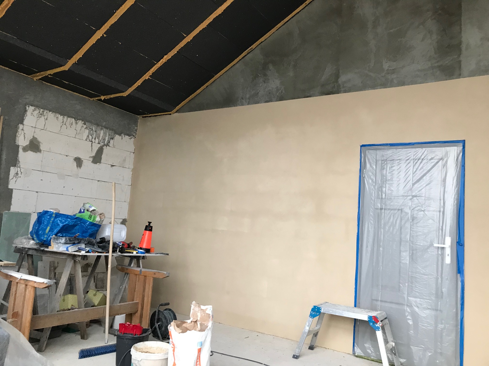 Wnętrze w trakcie prac wykończeniowych: gładź na ścianie, zabezpieczone drzwi folią malarską, widoczne narzędzia i materiały budowlane, sufit z izolacją.