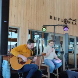 Duet muzyczny: mężczyzna gra na gitarze akustycznej w żółtej bluzie, kobieta śpiewa do mikrofonu trzymając kartkę, oświetlenie sceniczne w tle, drewniane ściany lokalu z napisem KUFONKA.