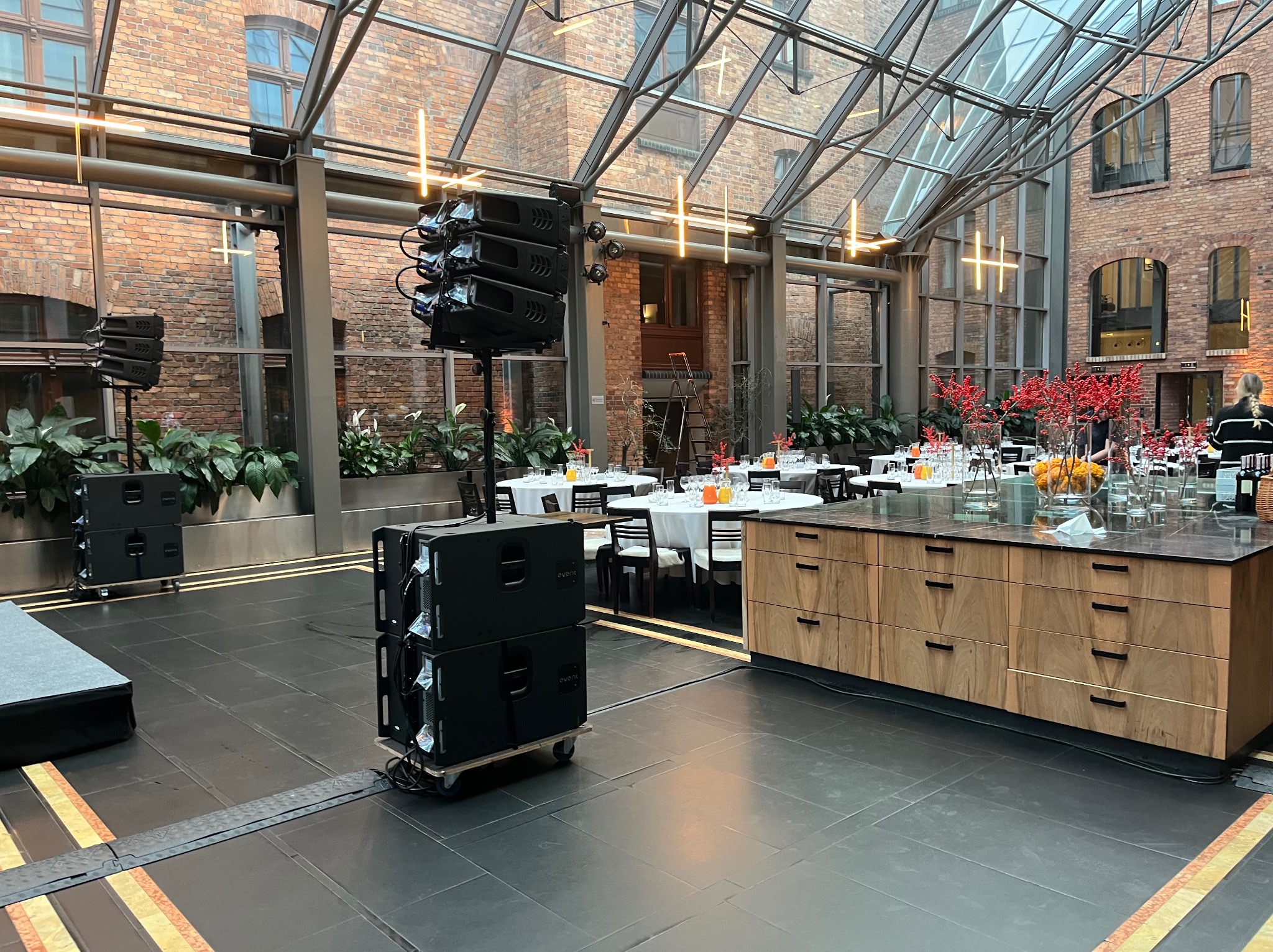 Sala bankietowa z przeszklonym dachem, przygotowana do imprezy, z nagłośnieniem na pierwszym planie i stołami z białymi obrusami w tle.