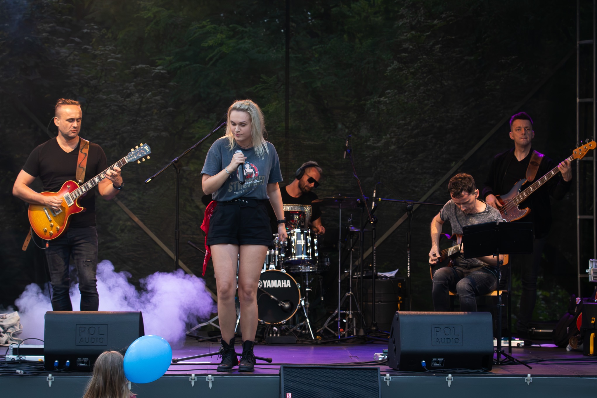 Zespół muzyczny gra na scenie podczas koncertu plenerowego, wokalistka śpiewa do mikrofonu, gitarzyści grają na gitarach elektrycznych, perkusista gra na perkusji Yamaha, widoczne nagłośnienie Pol...