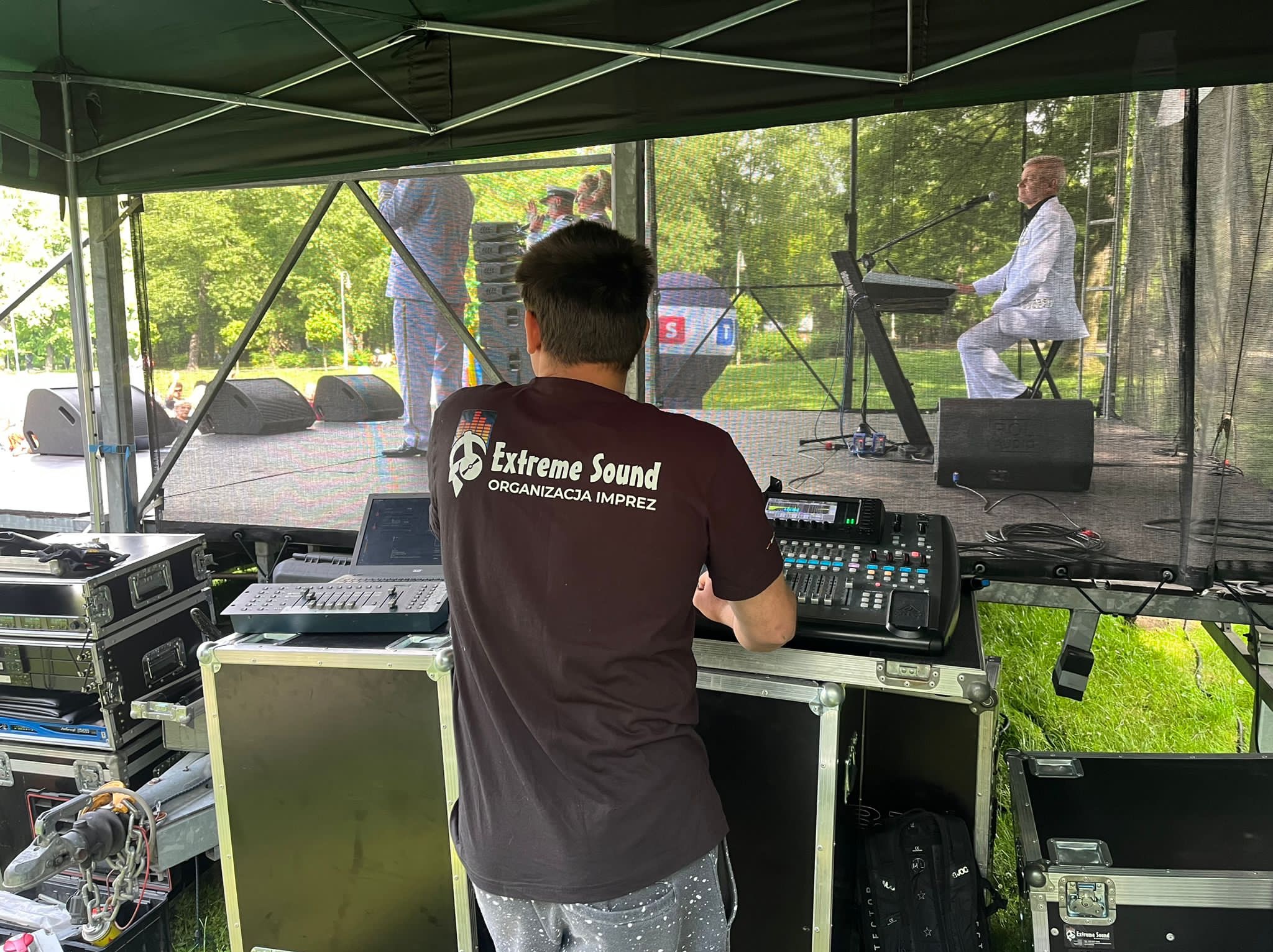 Osoba w koszulce firmy 'Extreme Sound Organizacja Imprez' obsługuje konsoletę dźwiękową podczas koncertu plenerowego, na scenie muzyk gra na keyboardzie.