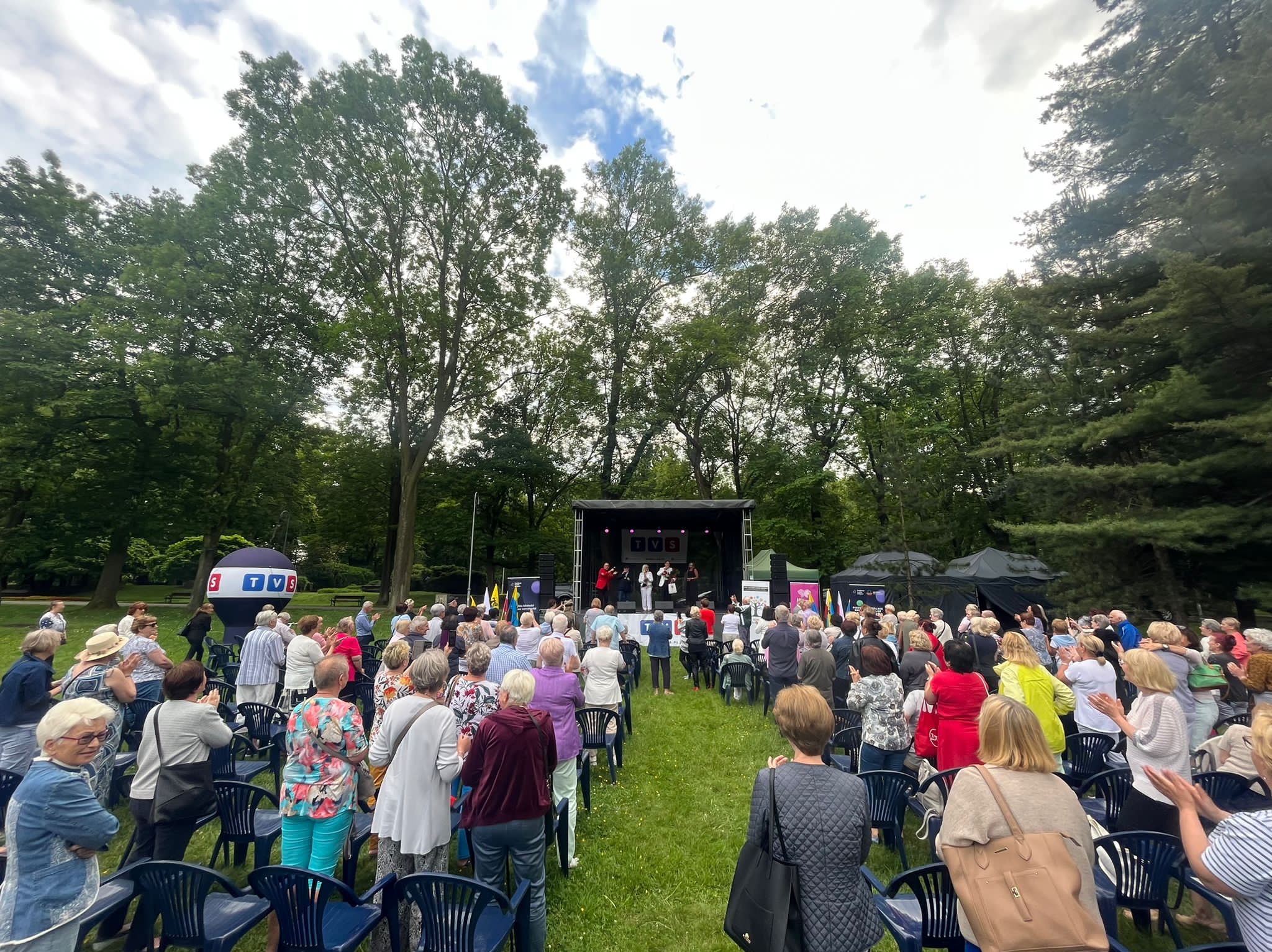 Plenerowa scena z występem na tle drzew, publiczność siedzi na krzesłach, w tle balon reklamowy TVS.