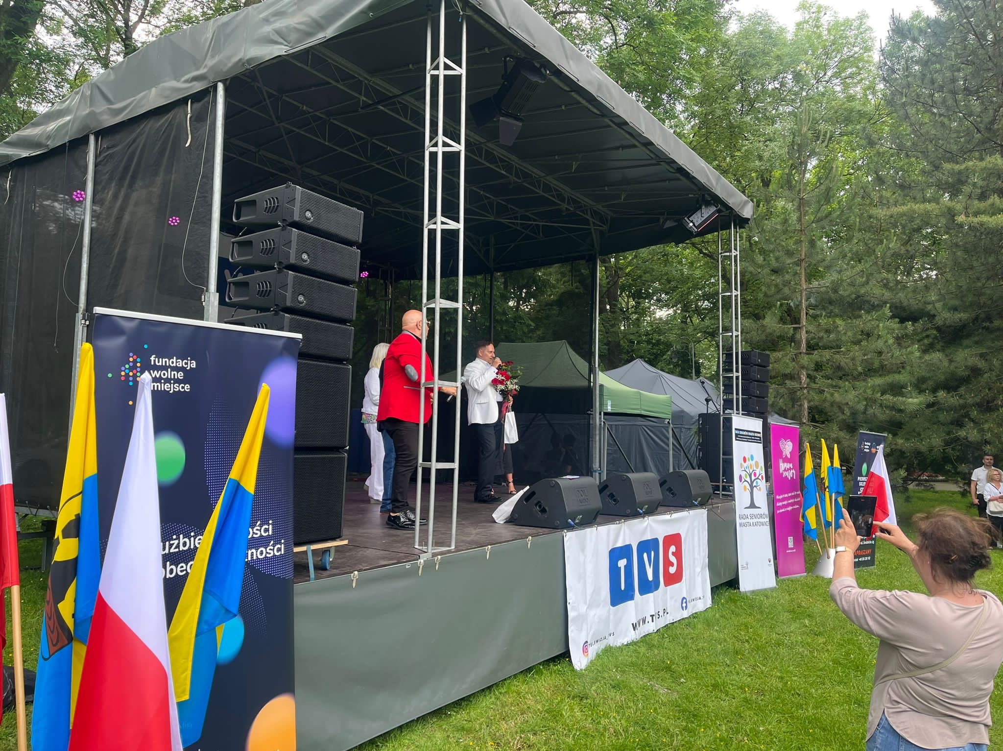 Scena plenerowa z nagłośnieniem i oświetleniem, na której przemawia mężczyzna z bukietem kwiatów, a obok stoi mężczyzna w czerwonej marynarce. W tle drzewa i transparenty. Osoba robi zdjęcie...