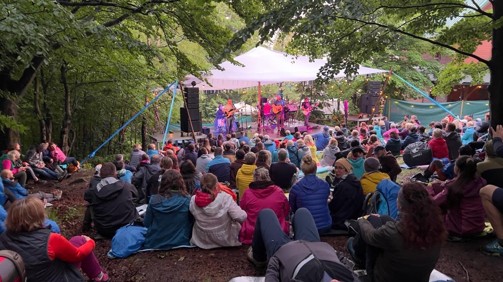 Plenerowy koncert w lesie: zespół na scenie pod białym namiotem gra dla zgromadzonego tłumu siedzącego na zboczu, widoczne drzewa i oświetlenie sceniczne.