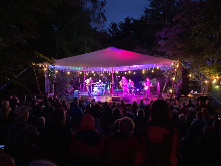 Zespół muzyczny gra koncert na zewnątrz pod namiotem oświetlonym girlandami żarówek, publiczność siedzi na trawie.