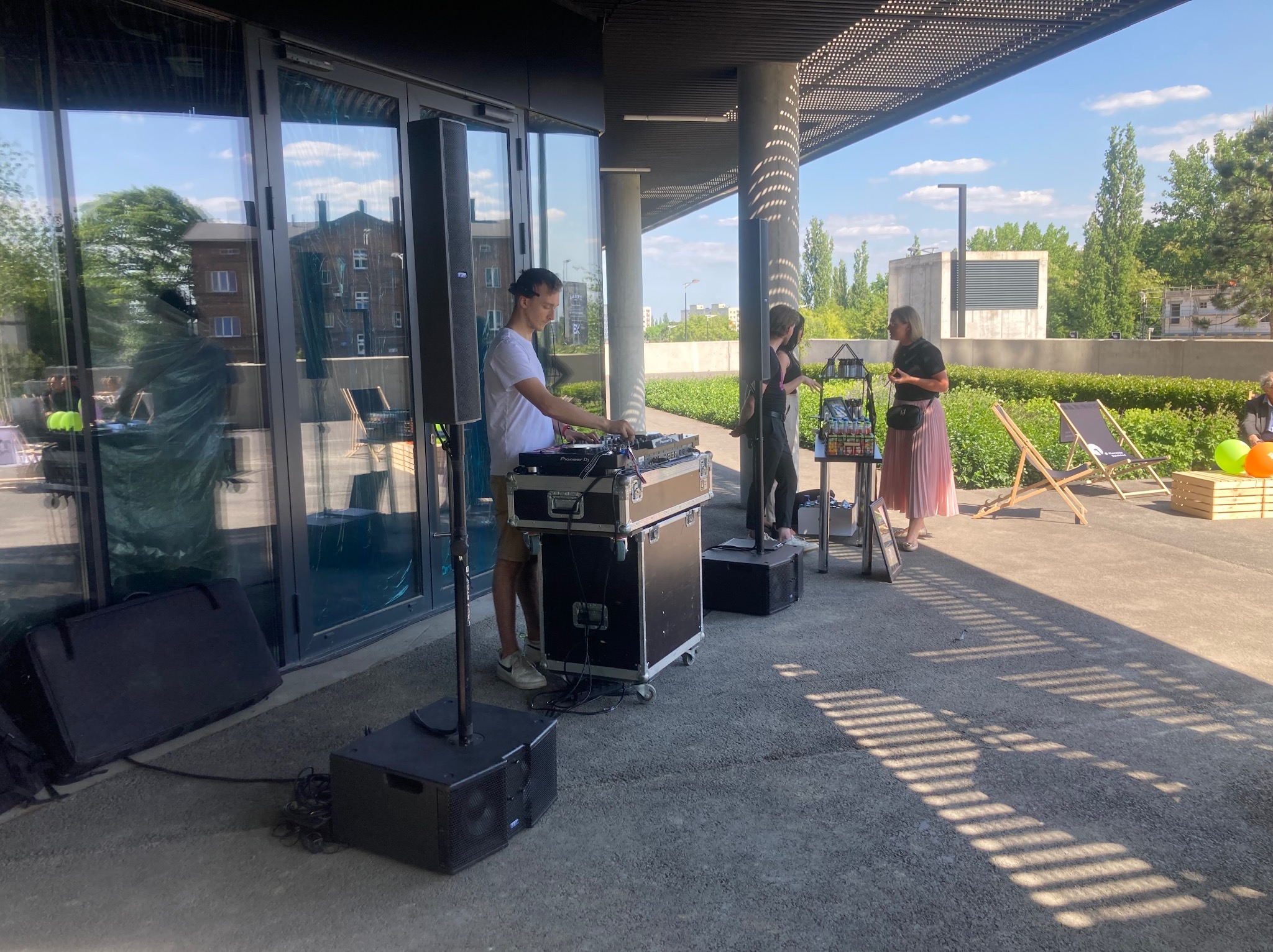 DJ miksuje muzykę na zewnątrz budynku, z głośnikami, sprzętem DJ-skim i osobami w tle, w słoneczny dzień.