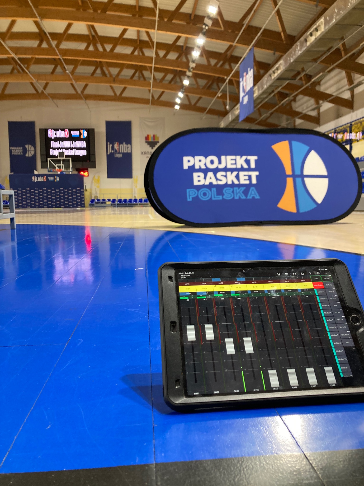 Tablet z aplikacją miksera audio na tle hali sportowej z banerem 'Projekt Basket Polska' podczas wydarzenia sportowego.