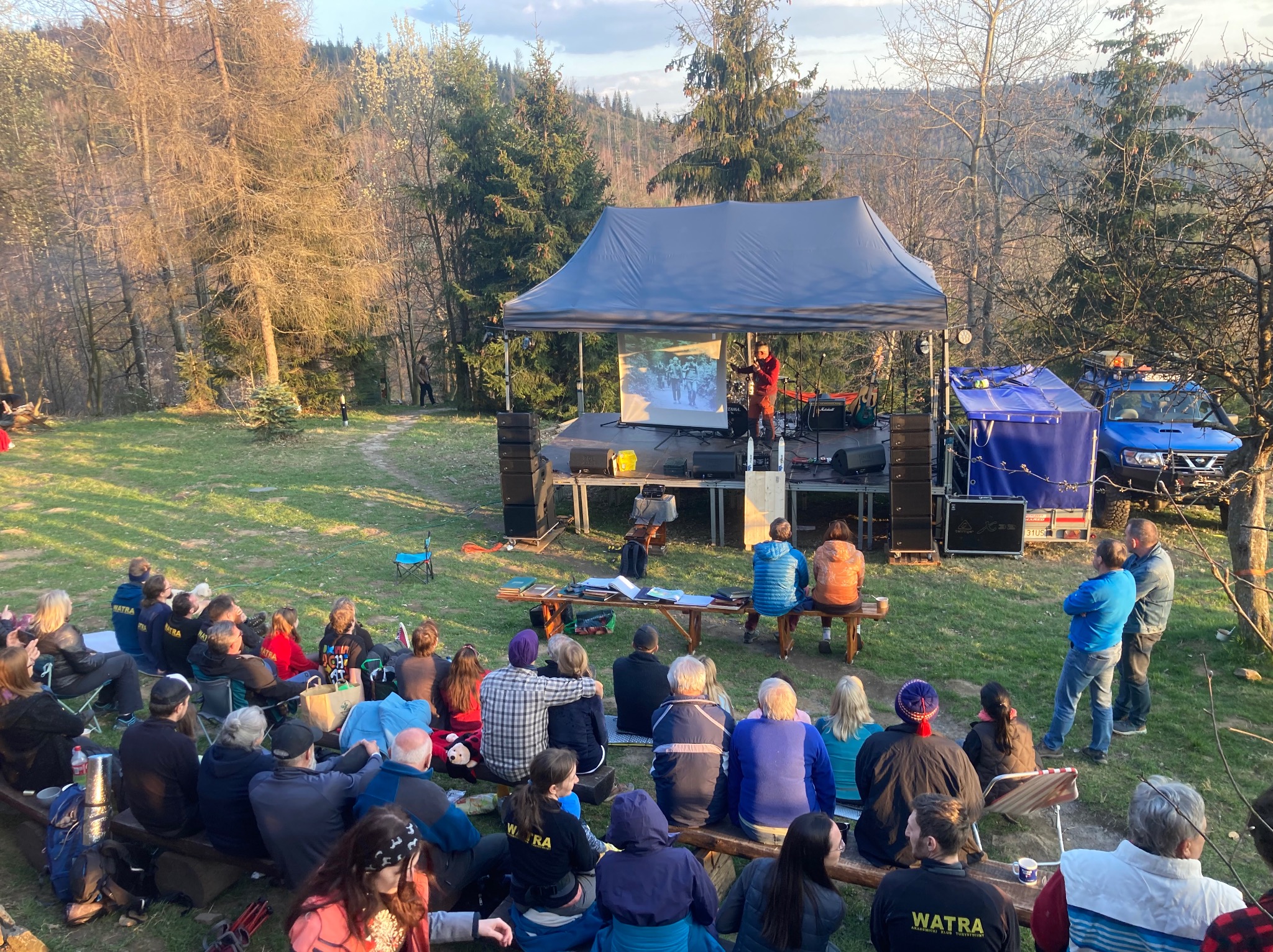 Plenerowa impreza integracyjna z występem na scenie i publicznością siedzącą na ławkach w otoczeniu drzew.