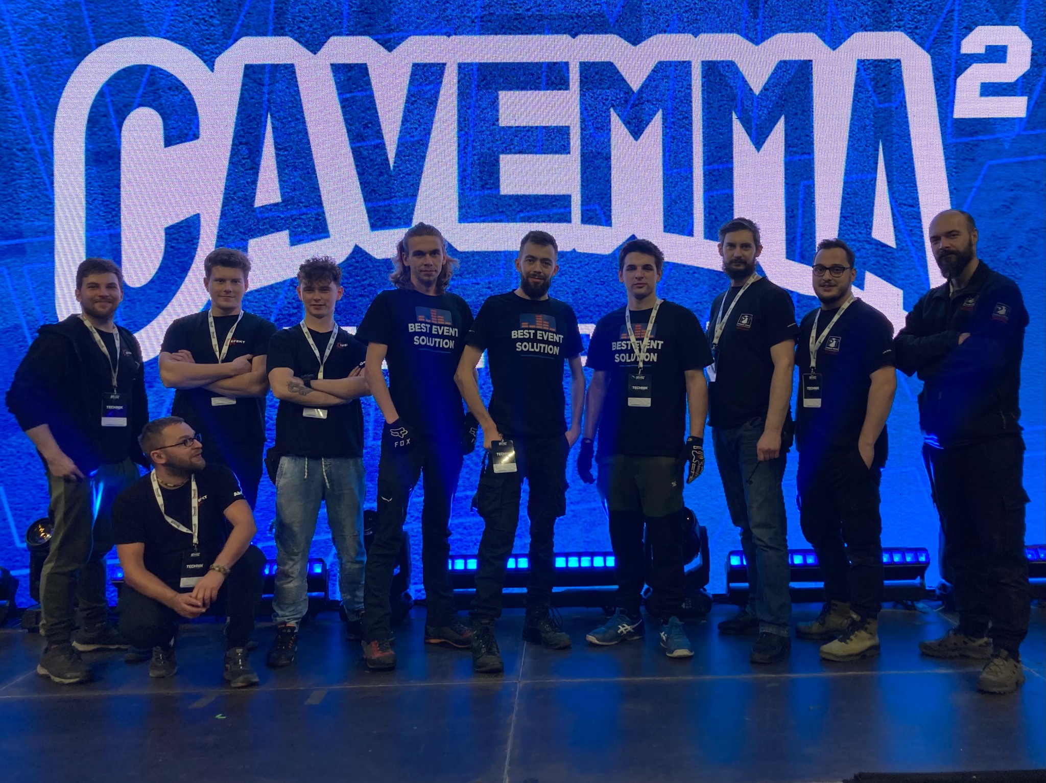 Grupa mężczyzn w czarnych koszulkach z napisem 'BEST EVENT SOLUTION' pozuje na tle podświetlonej ściany z dużym logo 'CAVEMMA 2', prawdopodobnie podczas wydarzenia.
