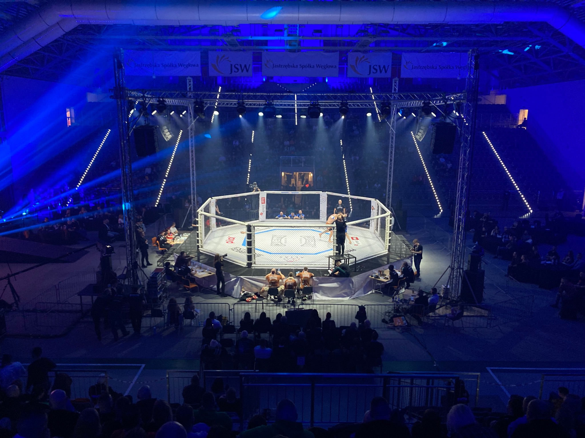 Widok z góry na arenę MMA oświetloną niebieskimi światłami, otoczoną publicznością i banerami sponsorskimi.