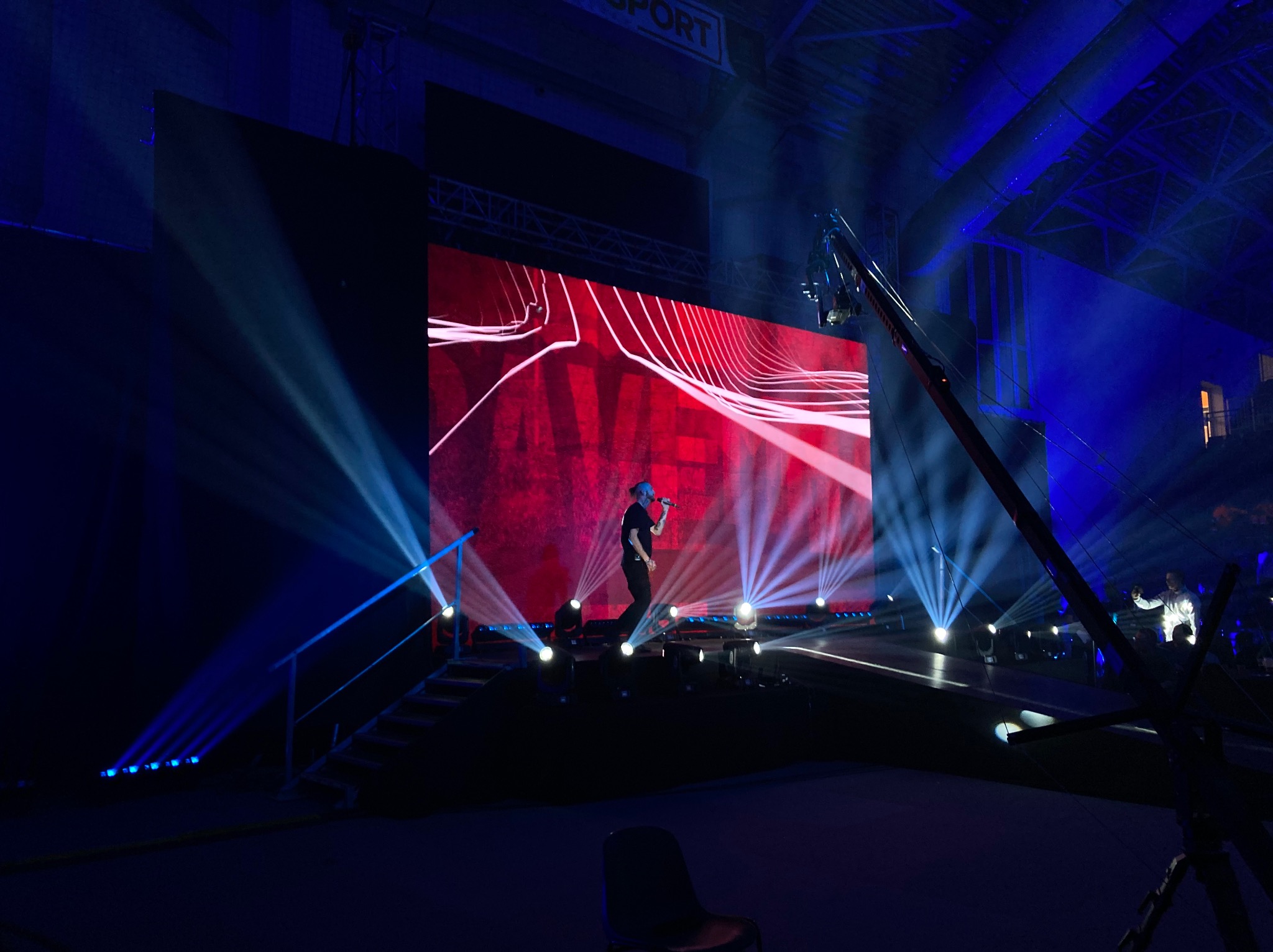 Występ artysty na scenie z dużym ekranem LED w tle, oświetlenie sceniczne w kolorze niebieskim, kamera na wysięgniku rejestruje wydarzenie.