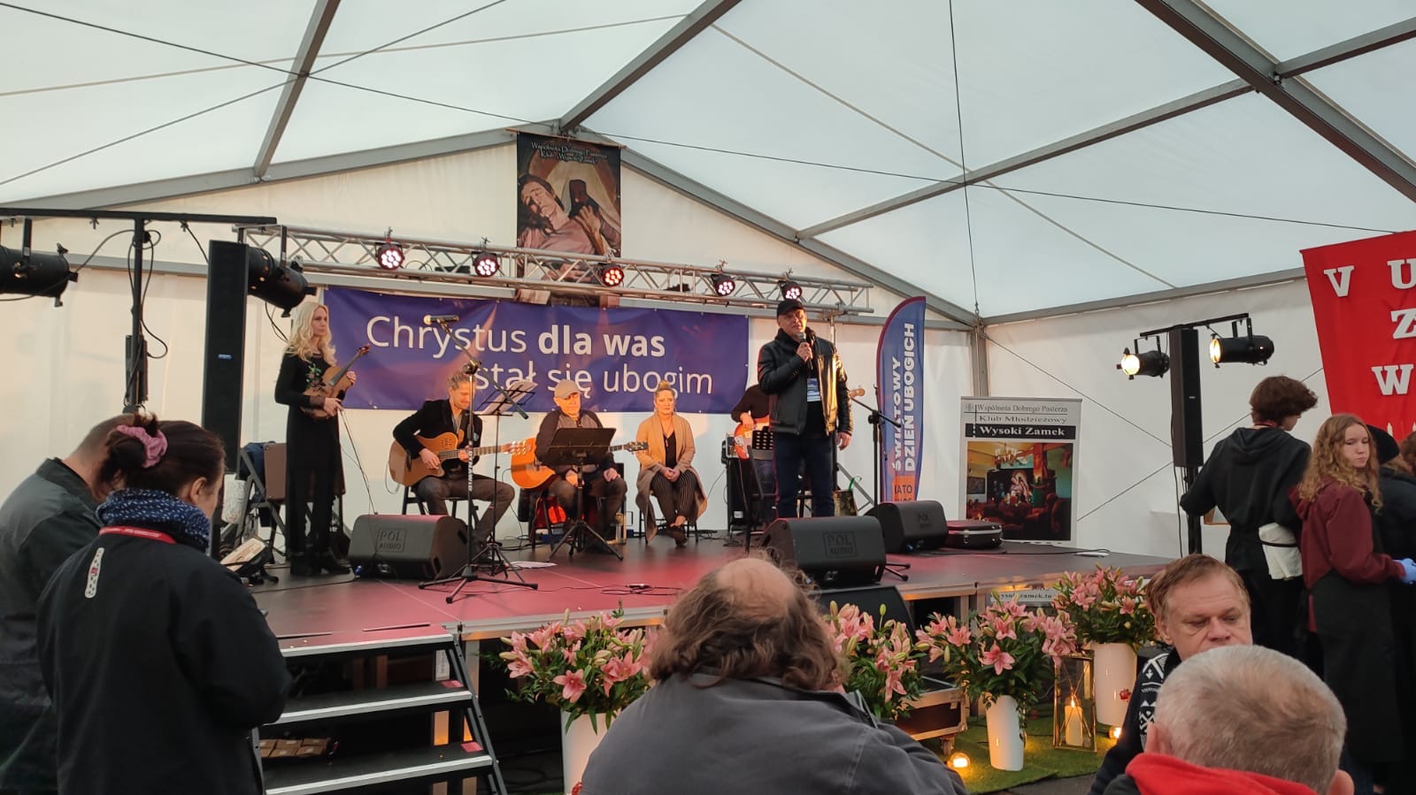 Zespół muzyczny i mówca na scenie pod namiotem podczas wydarzenia, widoczni widzowie, dekoracje z kwiatów.