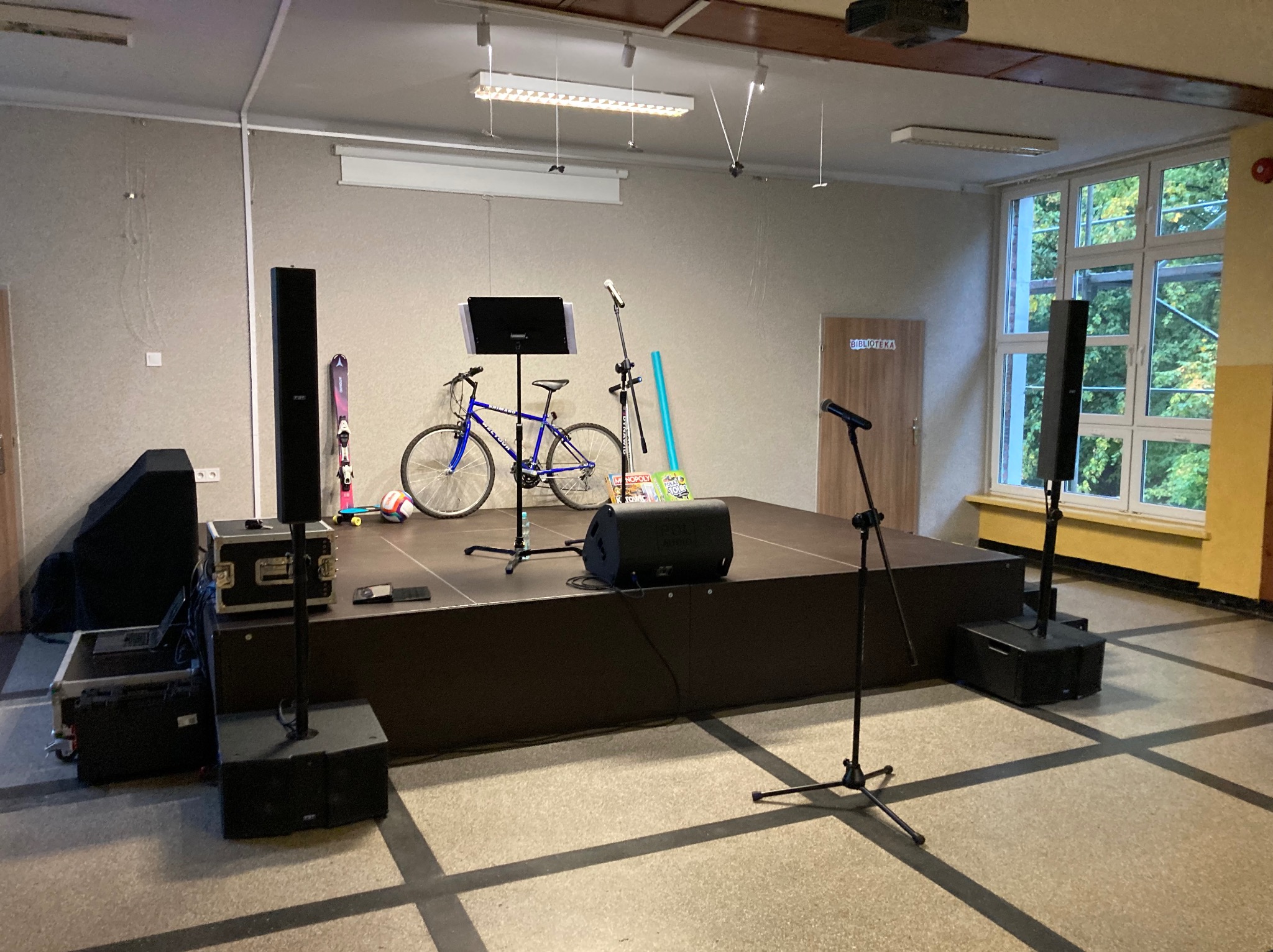 Scena z nagłośnieniem, mikrofonami i rowerem, przygotowana w sali z oknami i tablicą nutową.