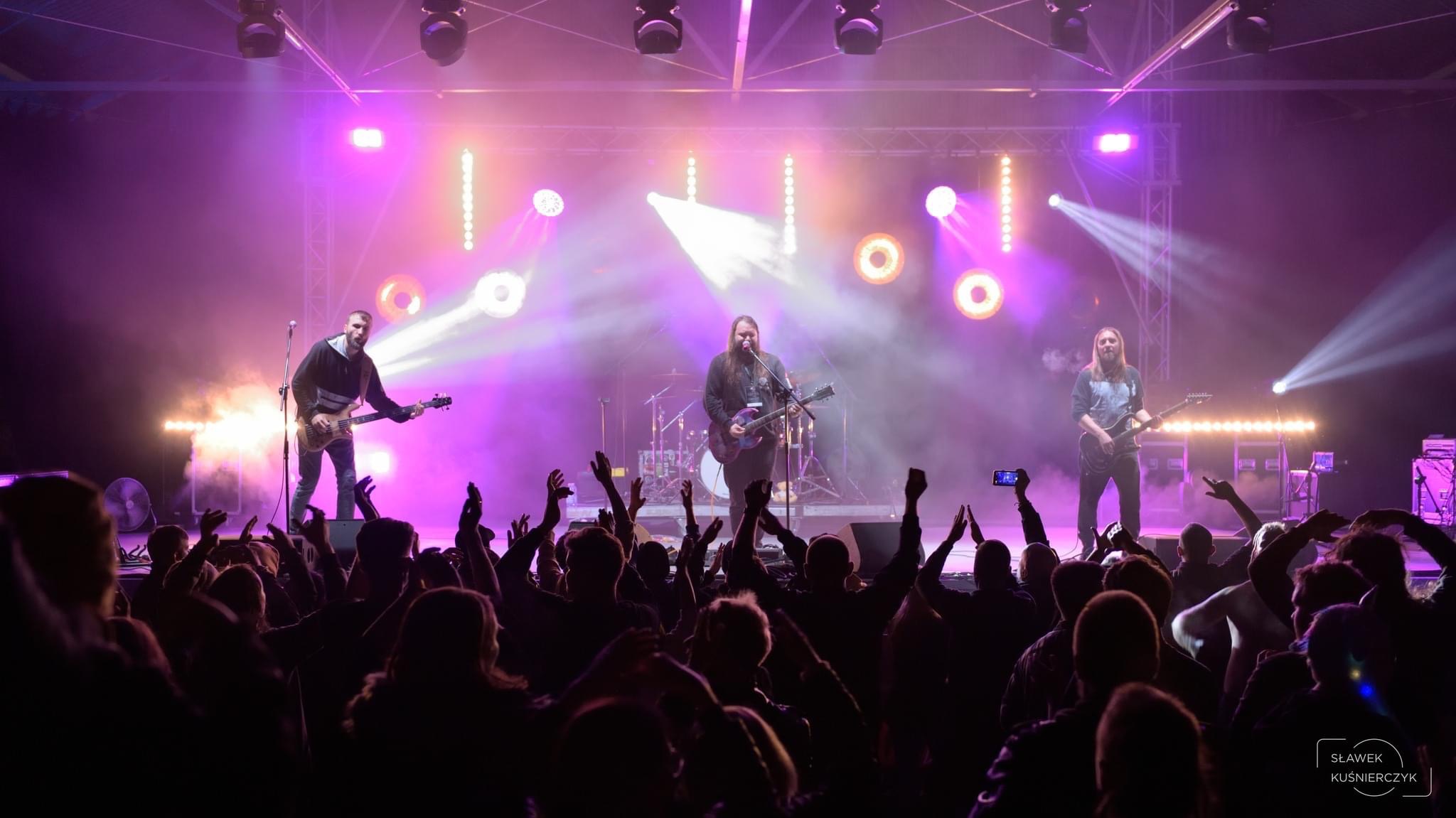 Zespół muzyczny na scenie w Katowicach, oświetlony fioletowymi światłami, grający dla publiczności z podniesionymi rękami.