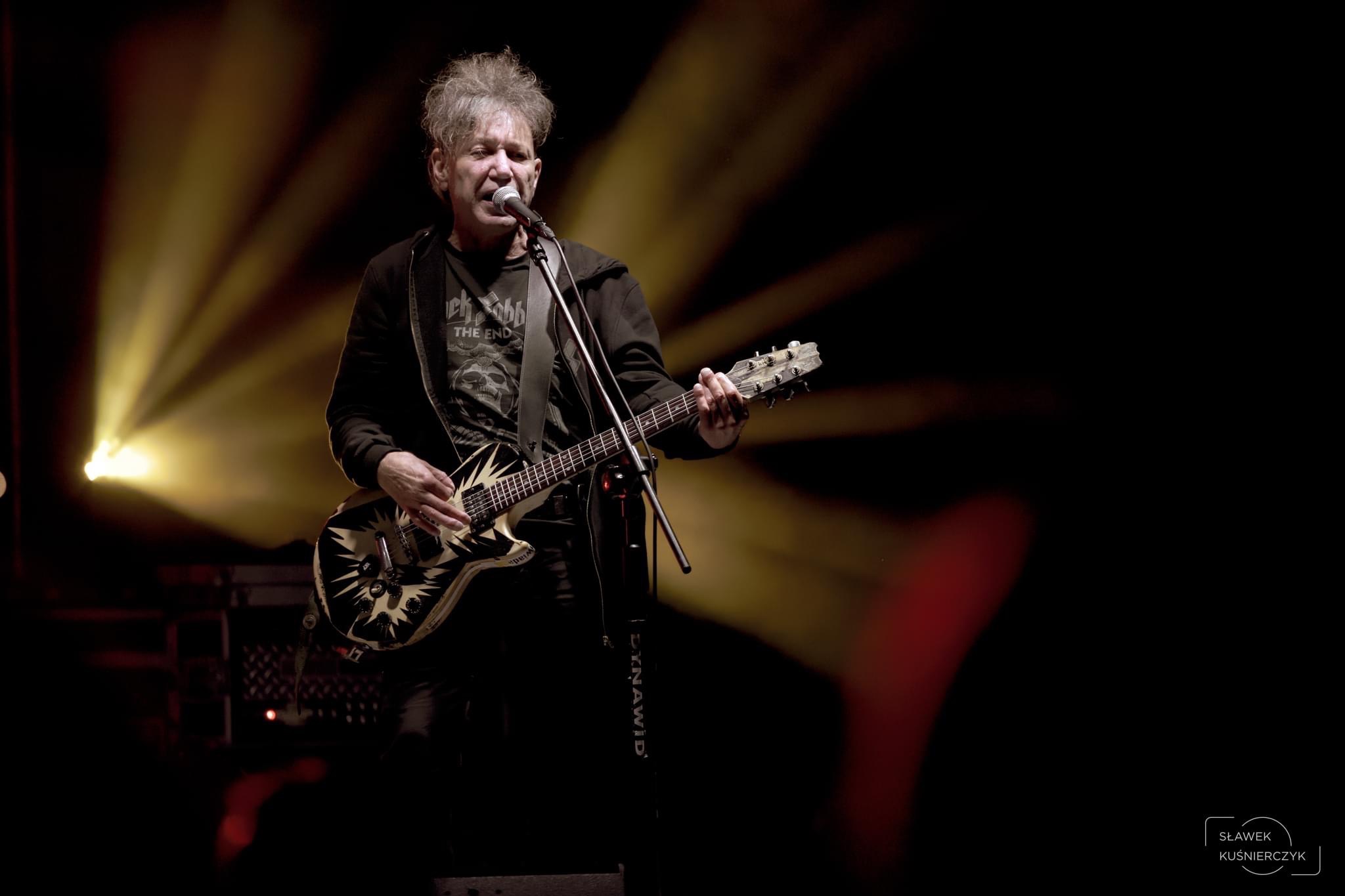 Mężczyzna z siwymi włosami śpiewa do mikrofonu i gra na elektrycznej gitarze podczas koncertu, oświetlony ciepłym światłem reflektorów.