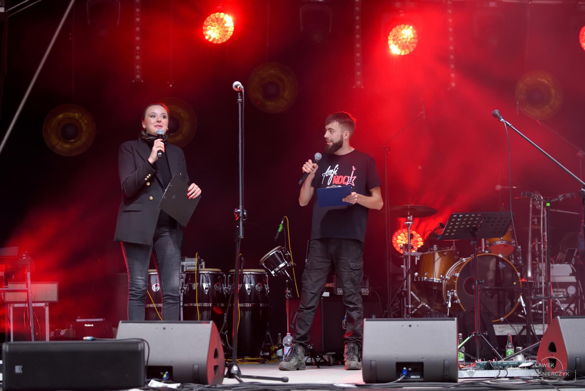 Dwoje konferansjerów na scenie oświetlonej czerwonymi światłami, za nimi zestaw perkusyjny i instrumenty muzyczne.