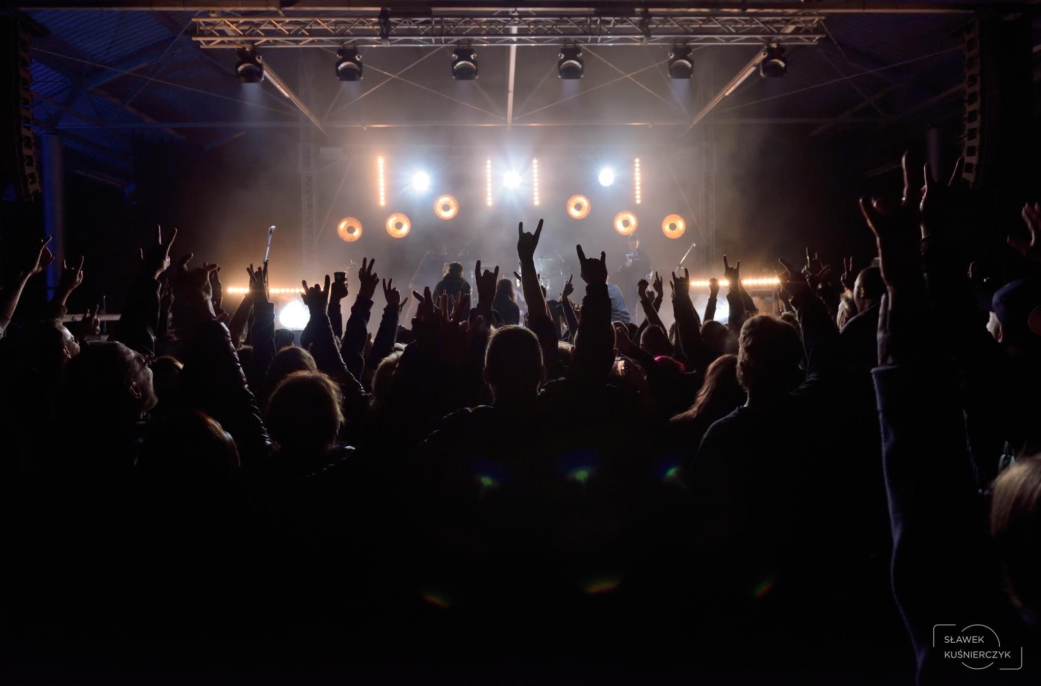 Koncert rockowy: Tłum fanów z podniesionymi rękami w geście 'diabła', scena oświetlona reflektorami, sylwetki muzyków w tle.
