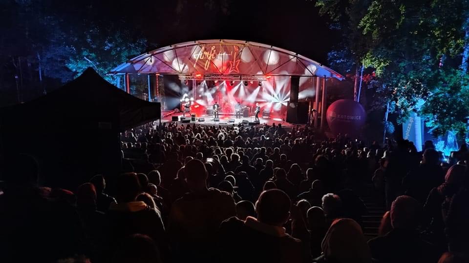 Widok na scenę oświetloną reflektorami podczas koncertu plenerowego, z tłumem widzów w ciemności i balonem z napisem Katowice w tle.