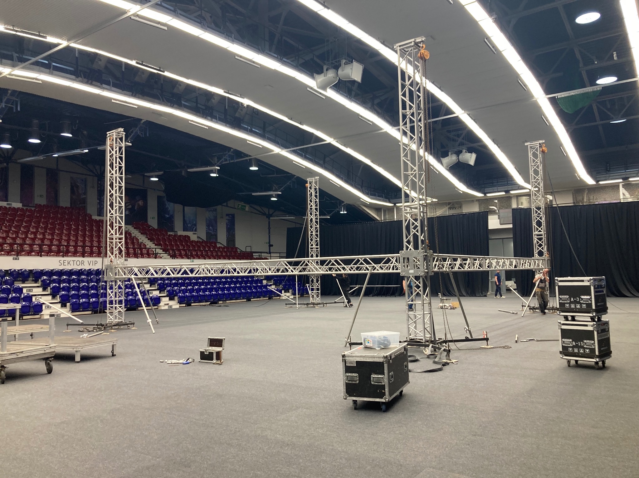 Montaż konstrukcji kratownicowej w hali widowiskowej, widoczne elementy oświetleniowe pod sufitem, krzesła dla publiczności w tle.