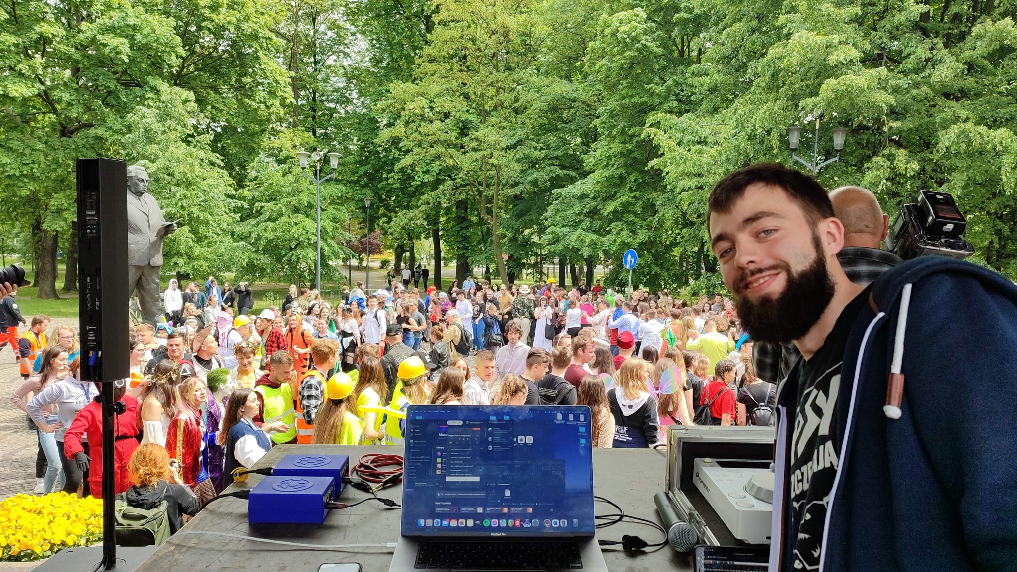 Mężczyzna uśmiecha się na tle imprezy plenerowej z nagłośnieniem, widoczny laptop z oprogramowaniem do miksowania muzyki, kolumna głośnikowa i statua.