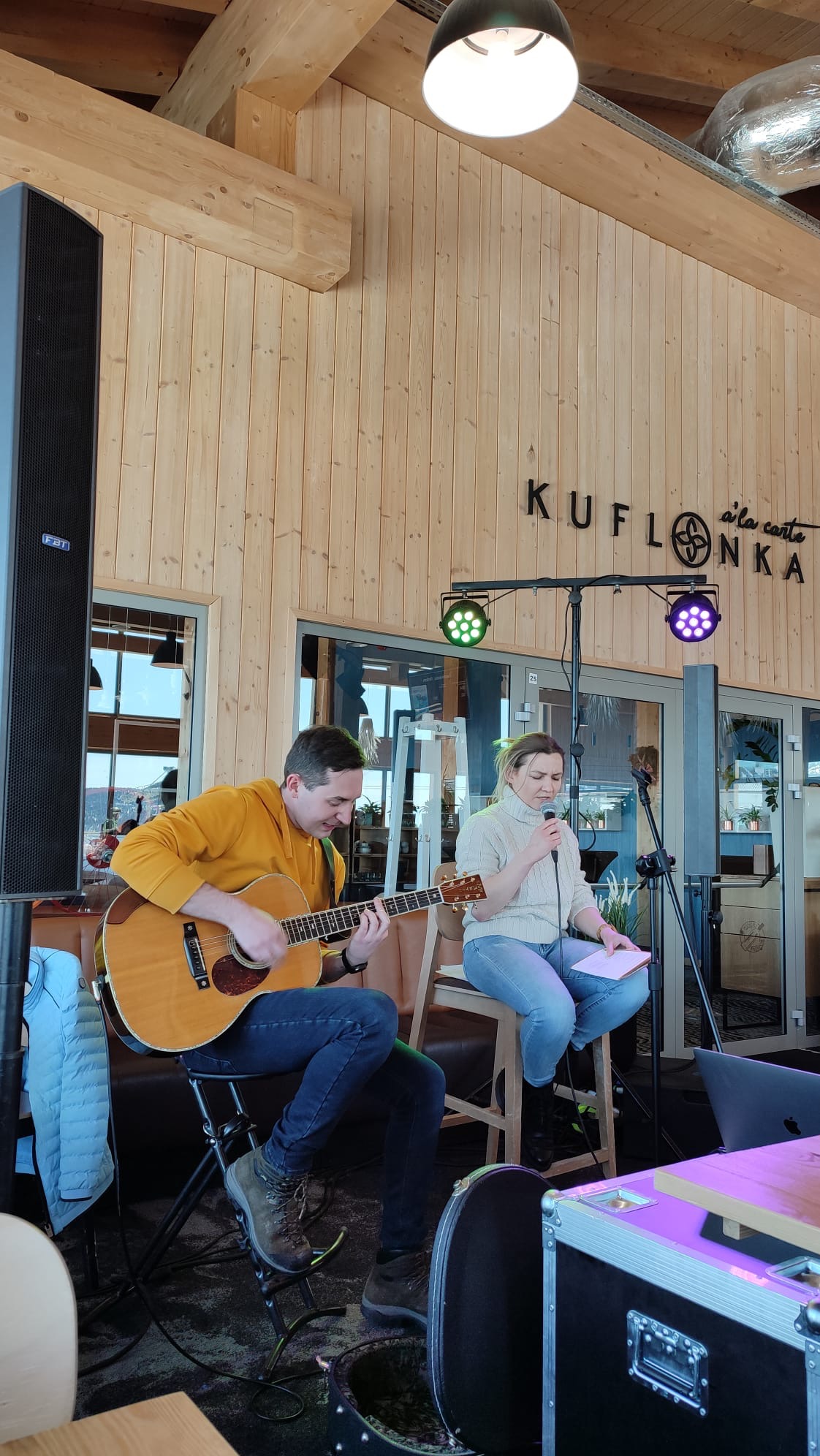 Duet muzyczny: mężczyzna gra na gitarze akustycznej w żółtej bluzie, kobieta śpiewa do mikrofonu trzymając kartkę, oświetlenie sceniczne w tle, drewniane ściany lokalu z napisem KUFONKA.