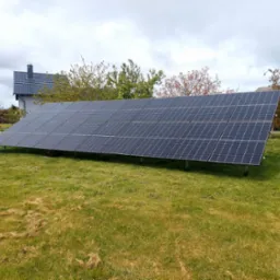 Panele fotowoltaiczne na trawniku przed domem w pochmurny dzień. Widoczne rzędy paneli ustawione na konstrukcji wsporczej. W tle dom z szarą dachówką.