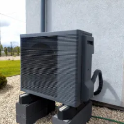 Szara pompa ciepła Panasonic Aquarea zamontowana na betonowych blokach przed jasnym budynkiem z kamiennym panelem, otoczona żwirowym podłożem i zielenią.