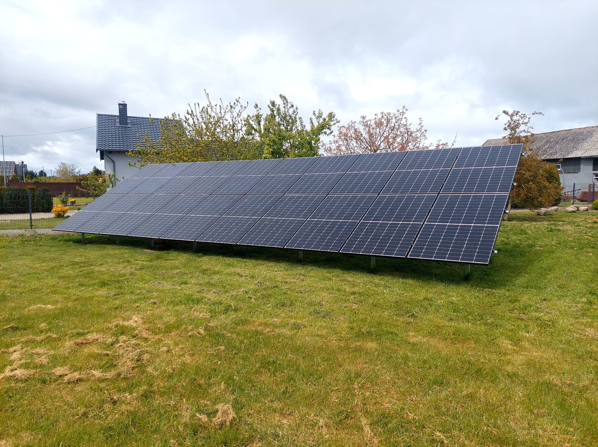 Panele fotowoltaiczne na trawniku przed domem w pochmurny dzień. Widoczne rzędy paneli ustawione na konstrukcji wsporczej. W tle dom z szarą dachówką.