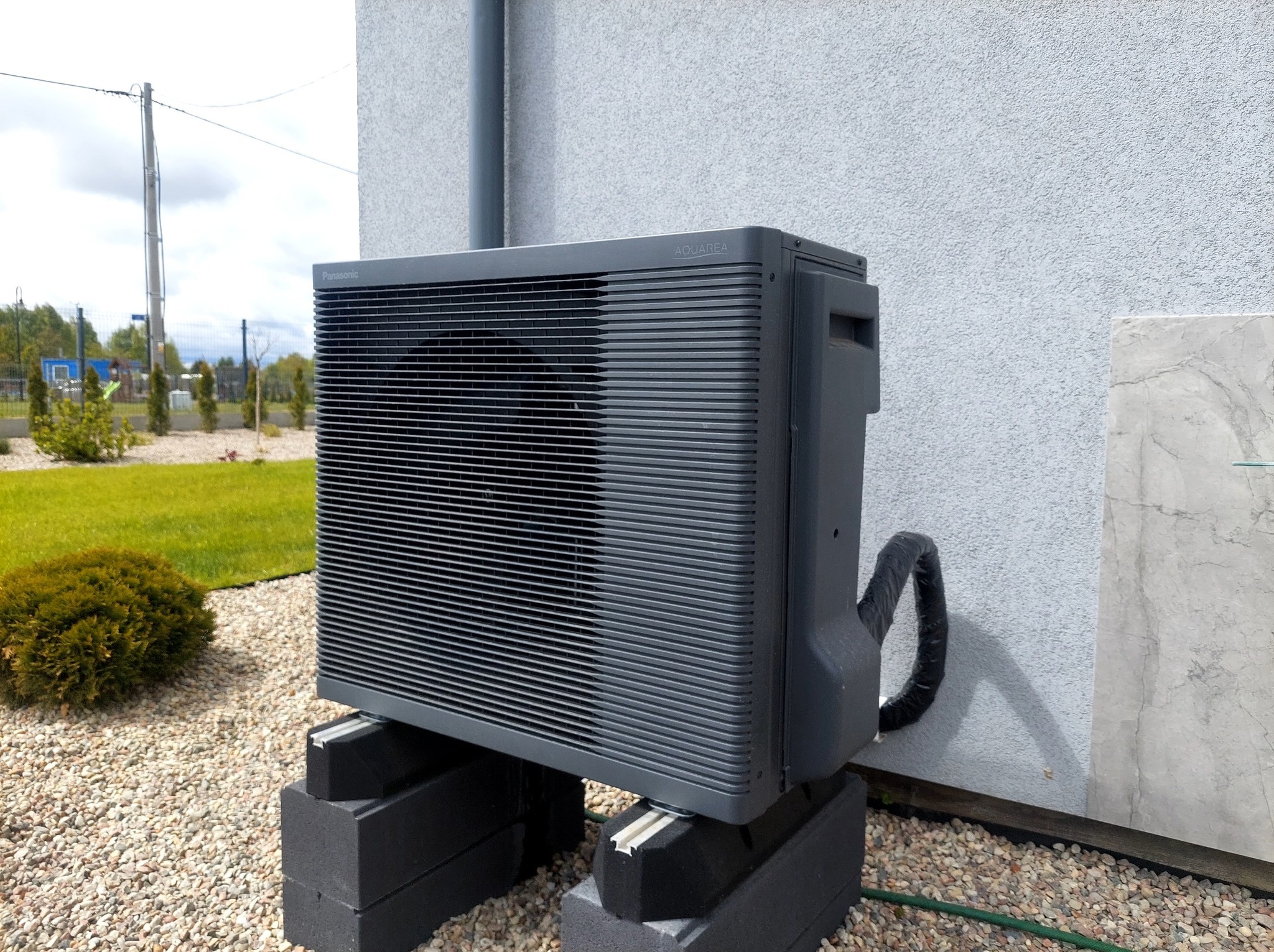 Szara pompa ciepła Panasonic Aquarea zamontowana na betonowych blokach przed jasnym budynkiem z kamiennym panelem, otoczona żwirowym podłożem i zielenią.