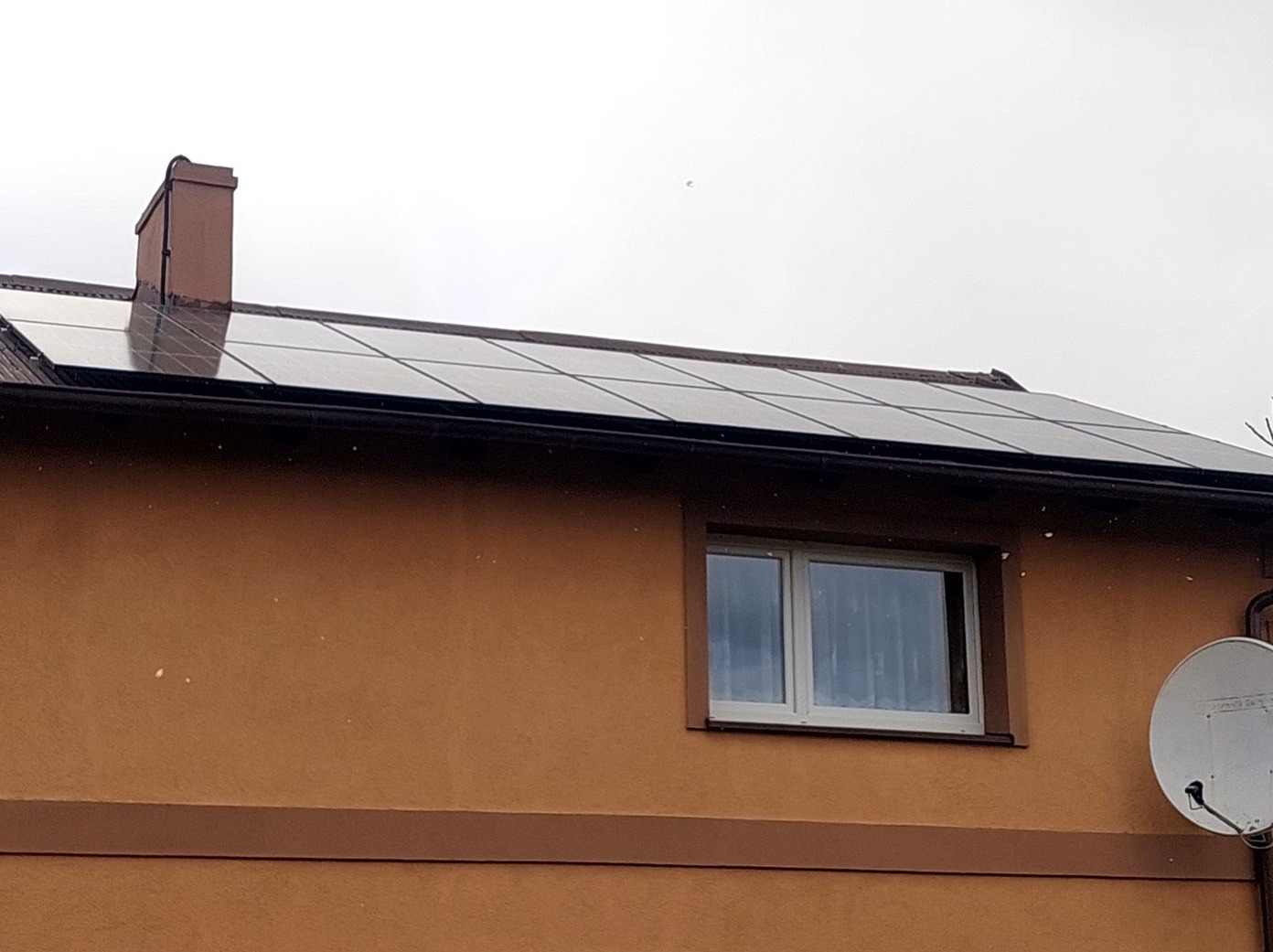 Panele fotowoltaiczne na dachu domu z brązową elewacją, widoczne okno i antena satelitarna. Pochmurne niebo w tle. Ujęcie z dołu, podkreślające instalację.
