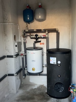 Zestaw kotłów z podłączonymi rurami w surowym pomieszczeniu. Biały i czarny zbiornik, dwa naczynia wzbiorcze. Widoczne zawory i manometry. Instalacja po montażu.